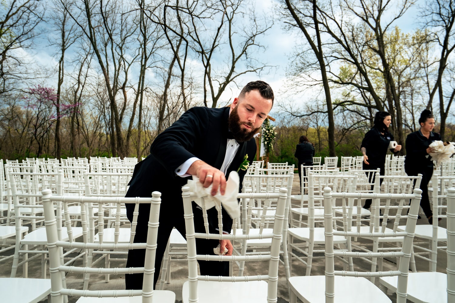 A candid picture of a groomsman wiping rainwater off Chivari chairs before an outside wedding ceremony at The Farmhouse KC Event Venue. 