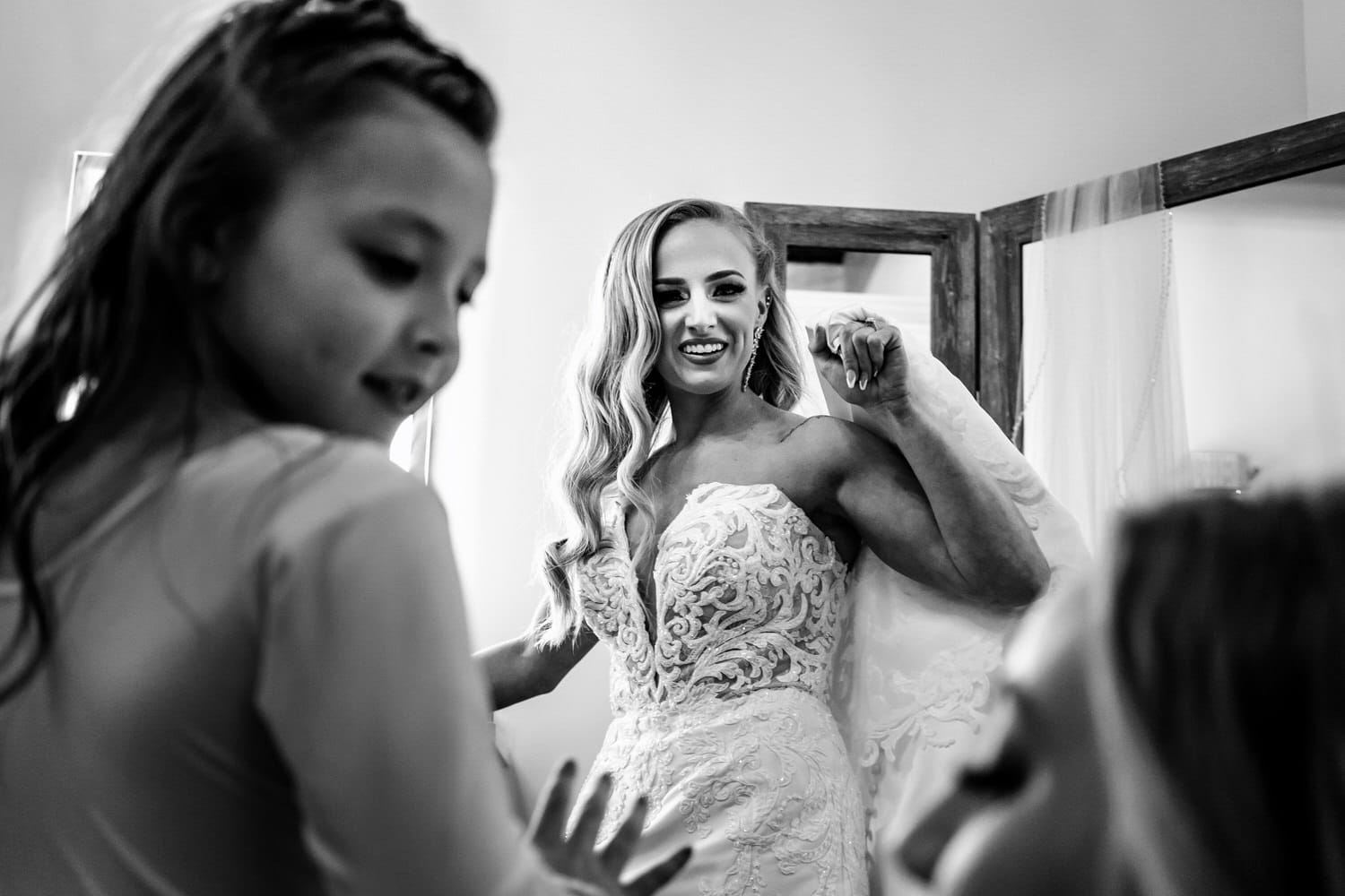 A candid black and white picture of a bride holding her wedding gown up as she watches a flower girl and bridesmaid on her spring wedding day. 
