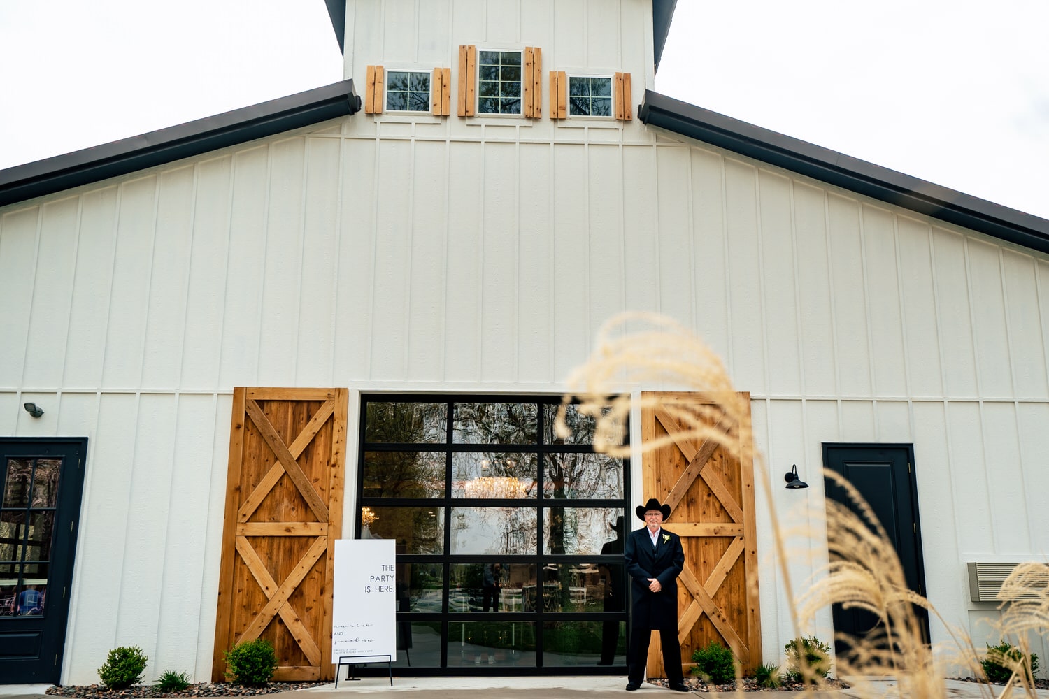 A wide, candid picture of a man in a tuxedo and black cowboy hat standing guard outside the bride's room at The Farmhouse KC Event Venue on a spring wedding day. 