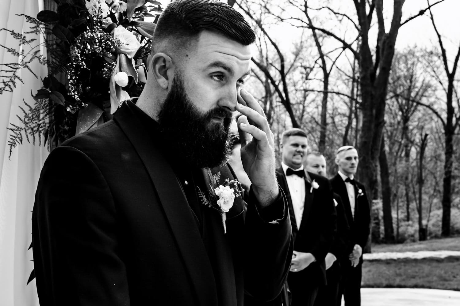 A candid black and white picture of a groom wiping tears from his eyes as he sees his bride for the first time during their spring wedding ceremony at The Farmhouse KC Event Venue. 