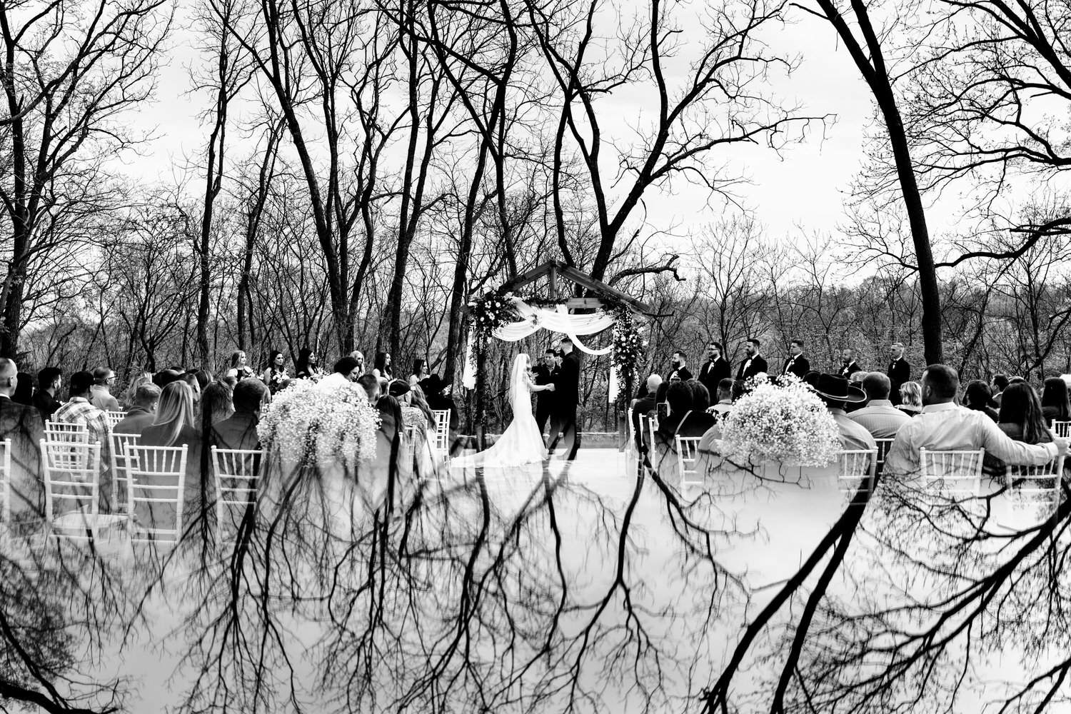 A wide, black and white picture of an outside wedding ceremony with the trees and sky reflected in the bottom half of the image at The Farmhouse KC Event Venue. 