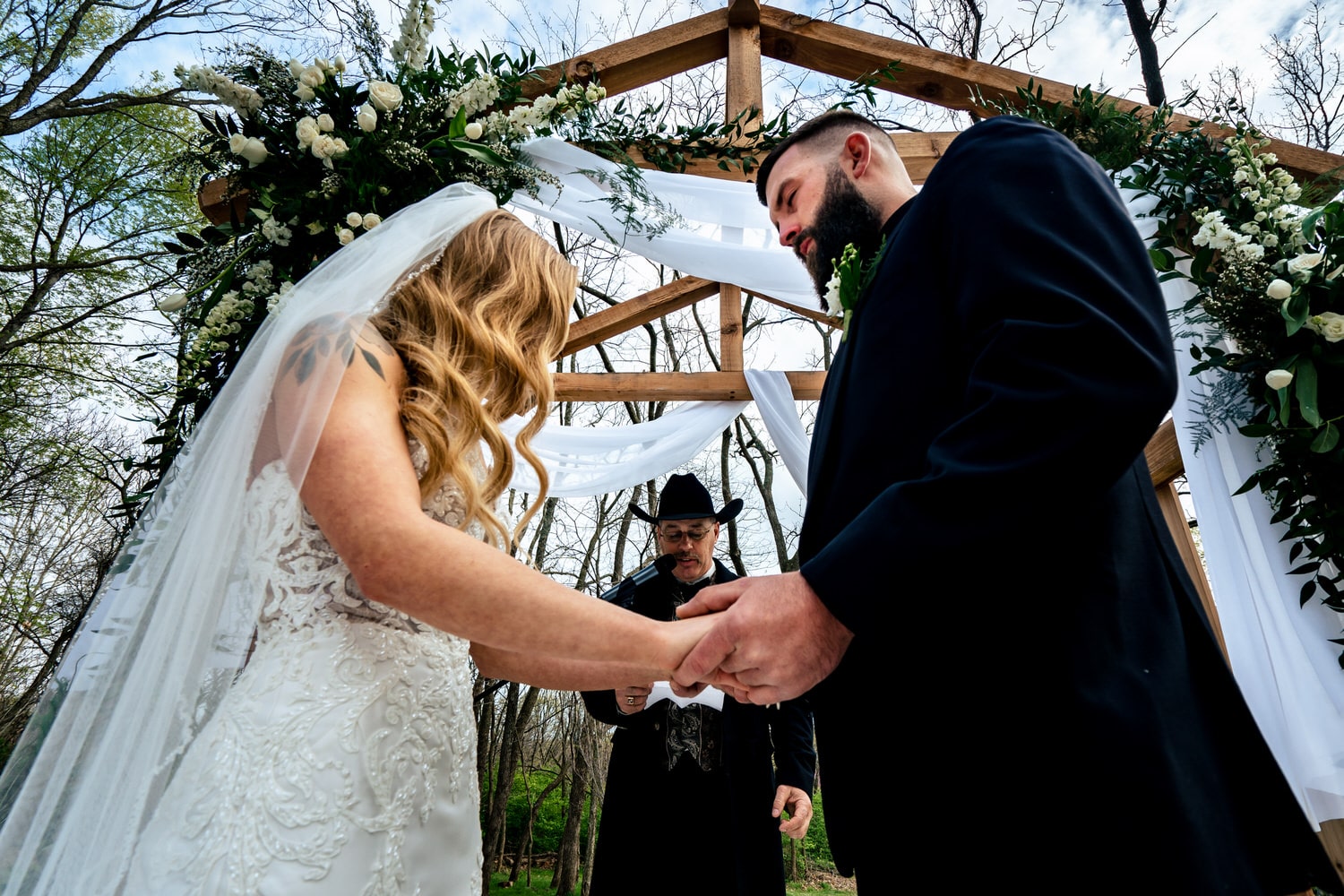 A candid, close-up picture of a bride and groom holding hands as they pray during their outside spring wedding ceremony at The Farmhouse KC Event Venue. 