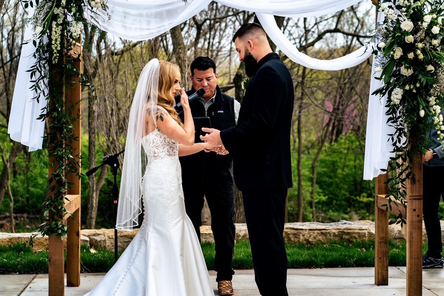 A candid, colorful picture of a bride wiping tears from her eyes as her groom puts a wedding band on her finger during an outside wedding ceremony at The Farmhouse KC Event Venue. 