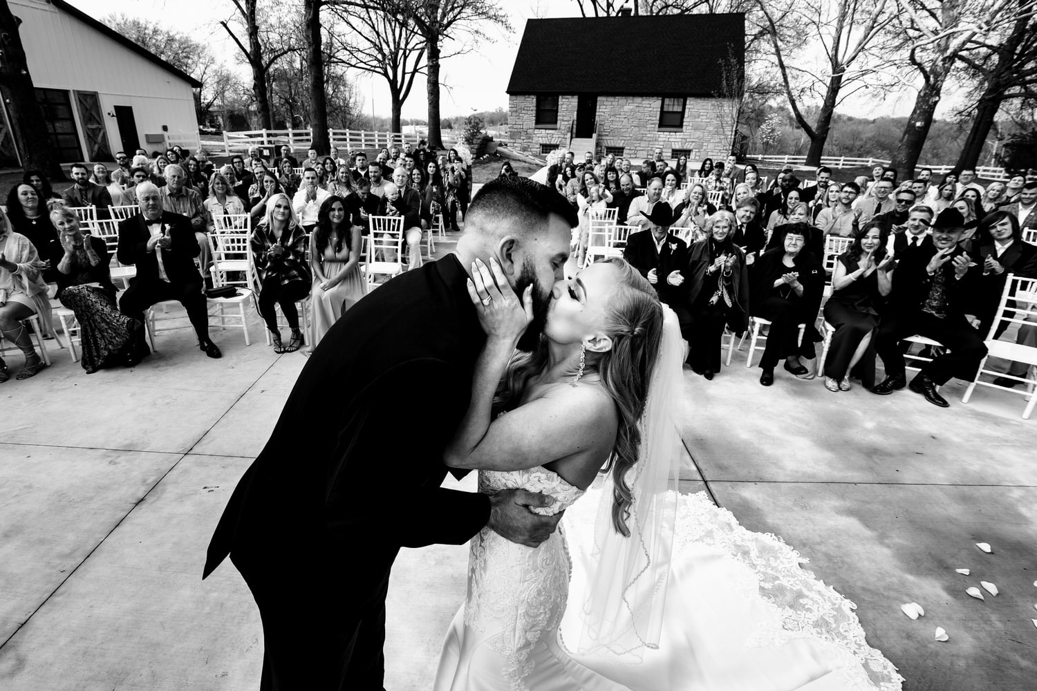 A candid black and white picture of a bride and groom sharing their first kiss, their family and friends visible in the background clapping and cheering. 