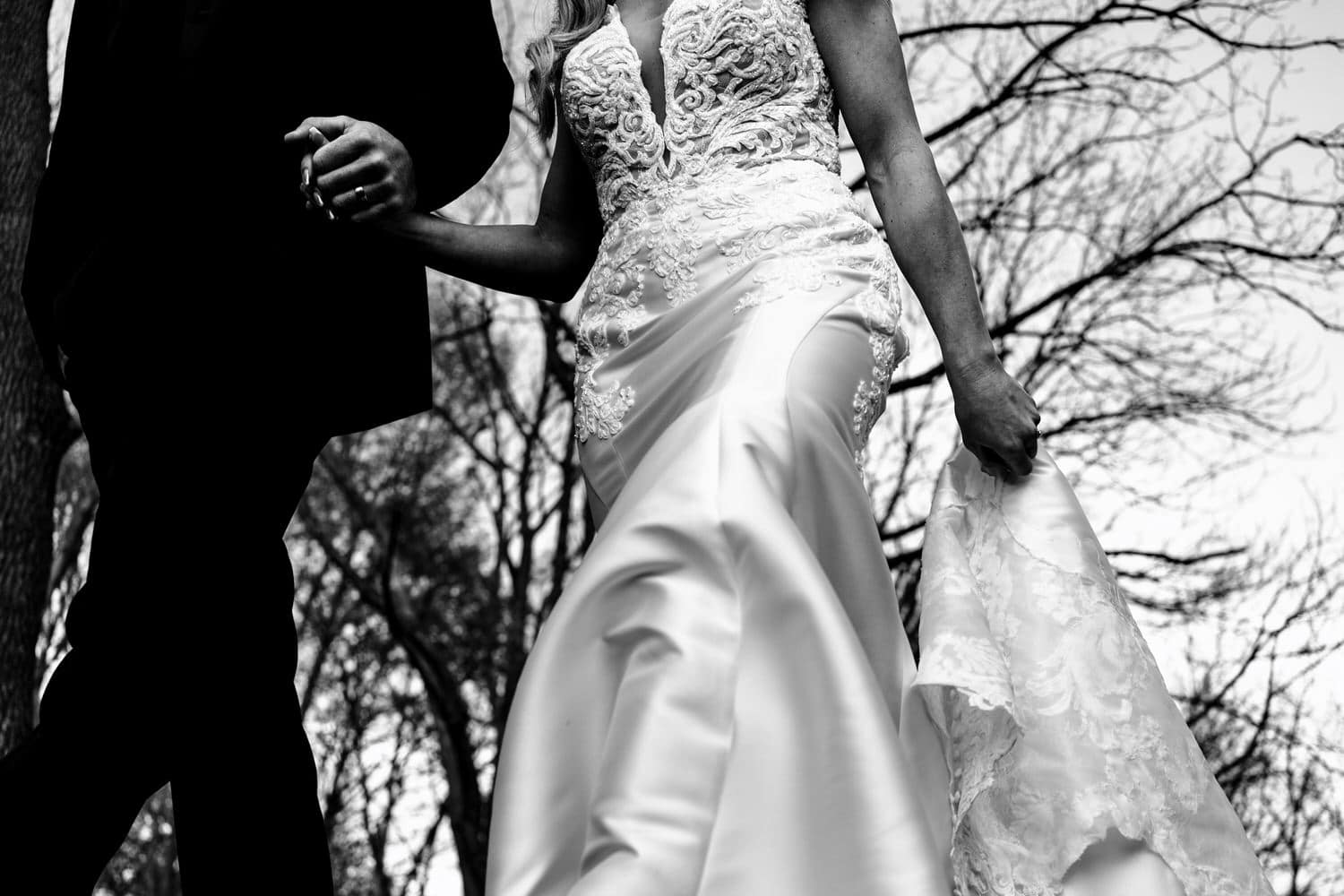 A close-up, black and white picture of a bride and groom holding hands, the bride's other hand holding up the train of her wedding gown. 