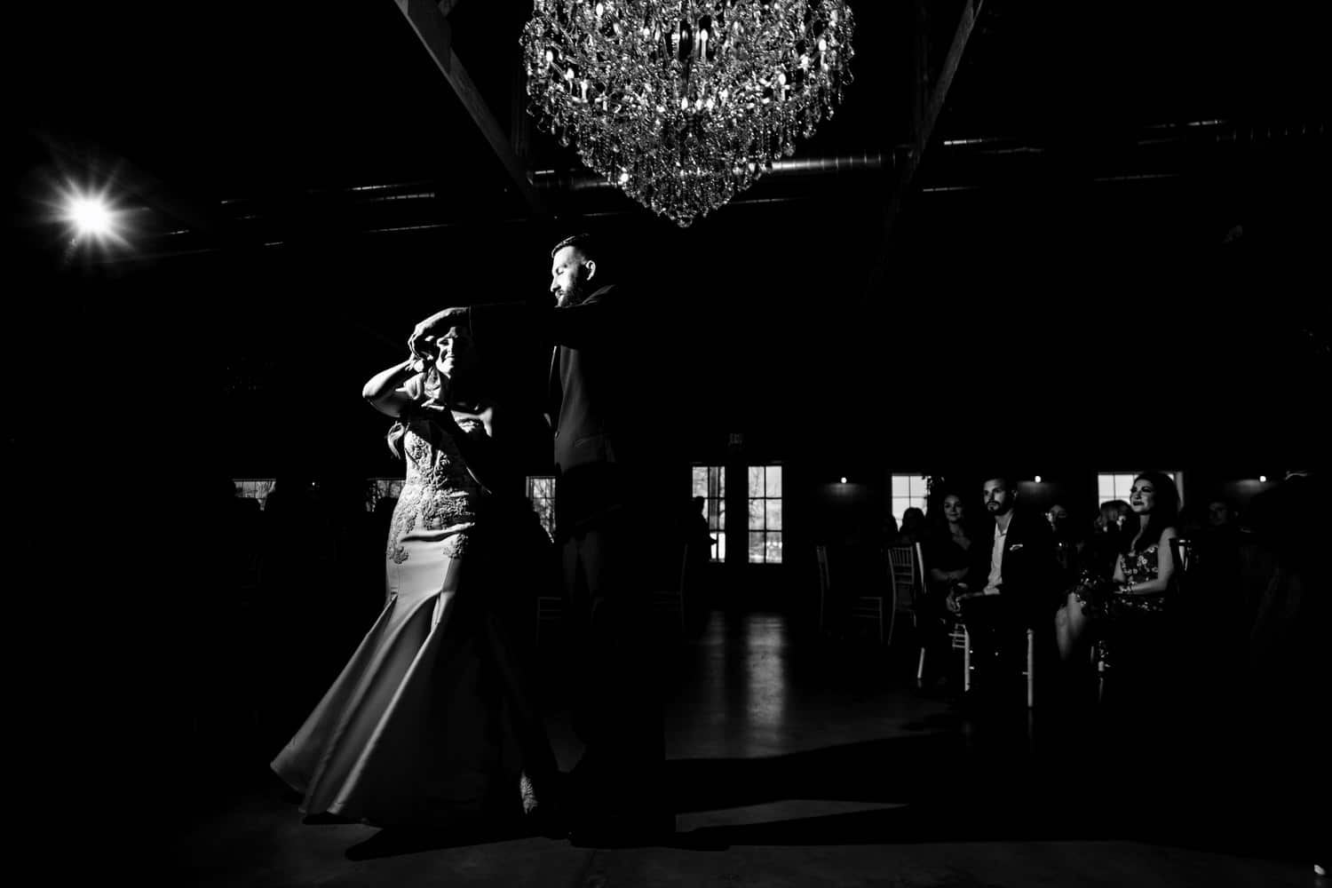 A candid black and white picture of a groom twirling his bride underneath his arm during their first dance at their spring wedding reception at The Farmhouse KC Event Venue. 