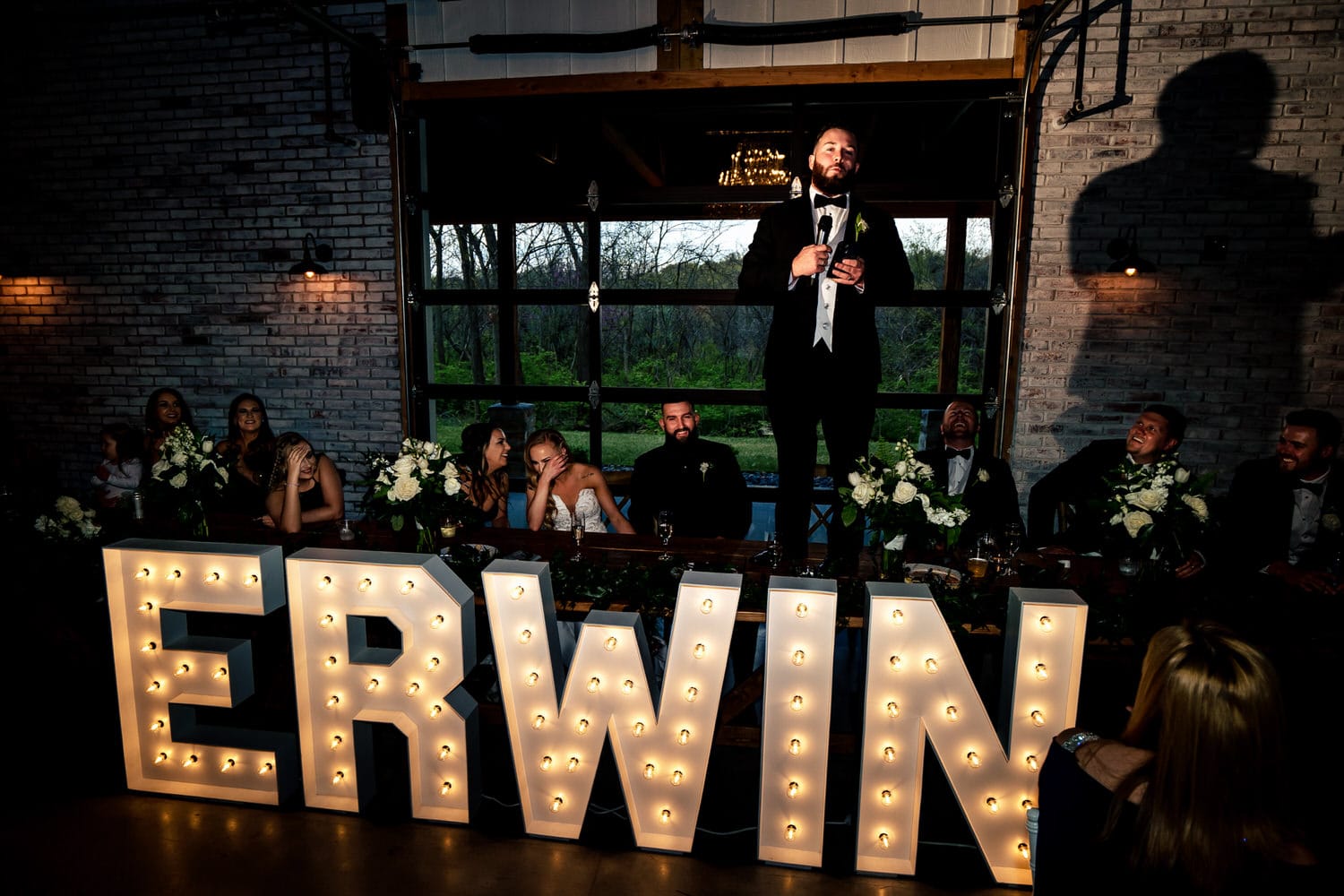 A candid picture of a best man standing on a table, giving a speech as a bride and groom laugh during their wedding reception at The Farmhouse KC Event Venue. 