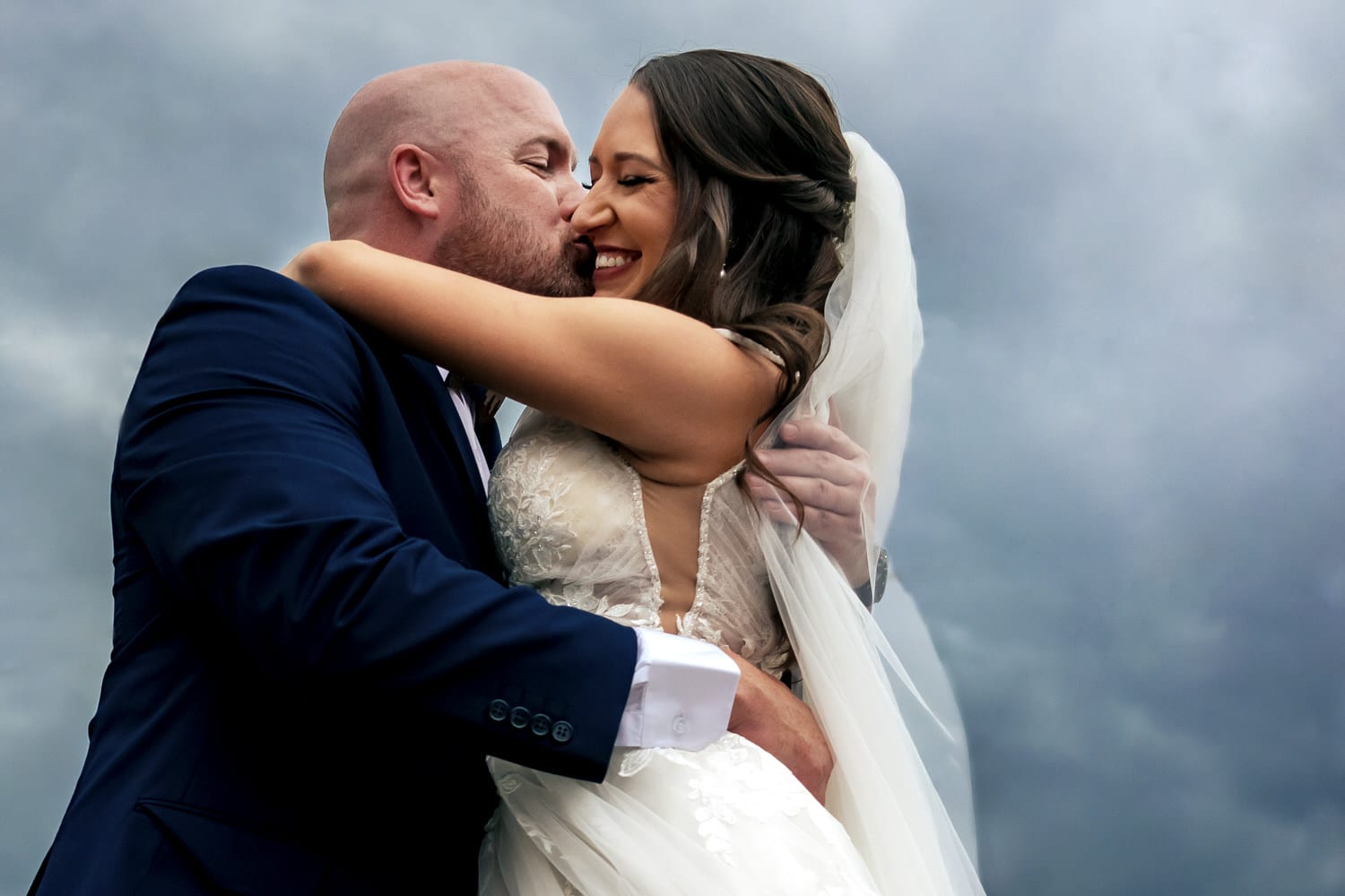 A colorful, candid picture of a groom in a blue tuxedo, hugging his bride tightly and kissing her cheek as she smiles widely against a dark and stormy sky on the day of their summer wedding at The Bride and The Bauer in Kansas City. 