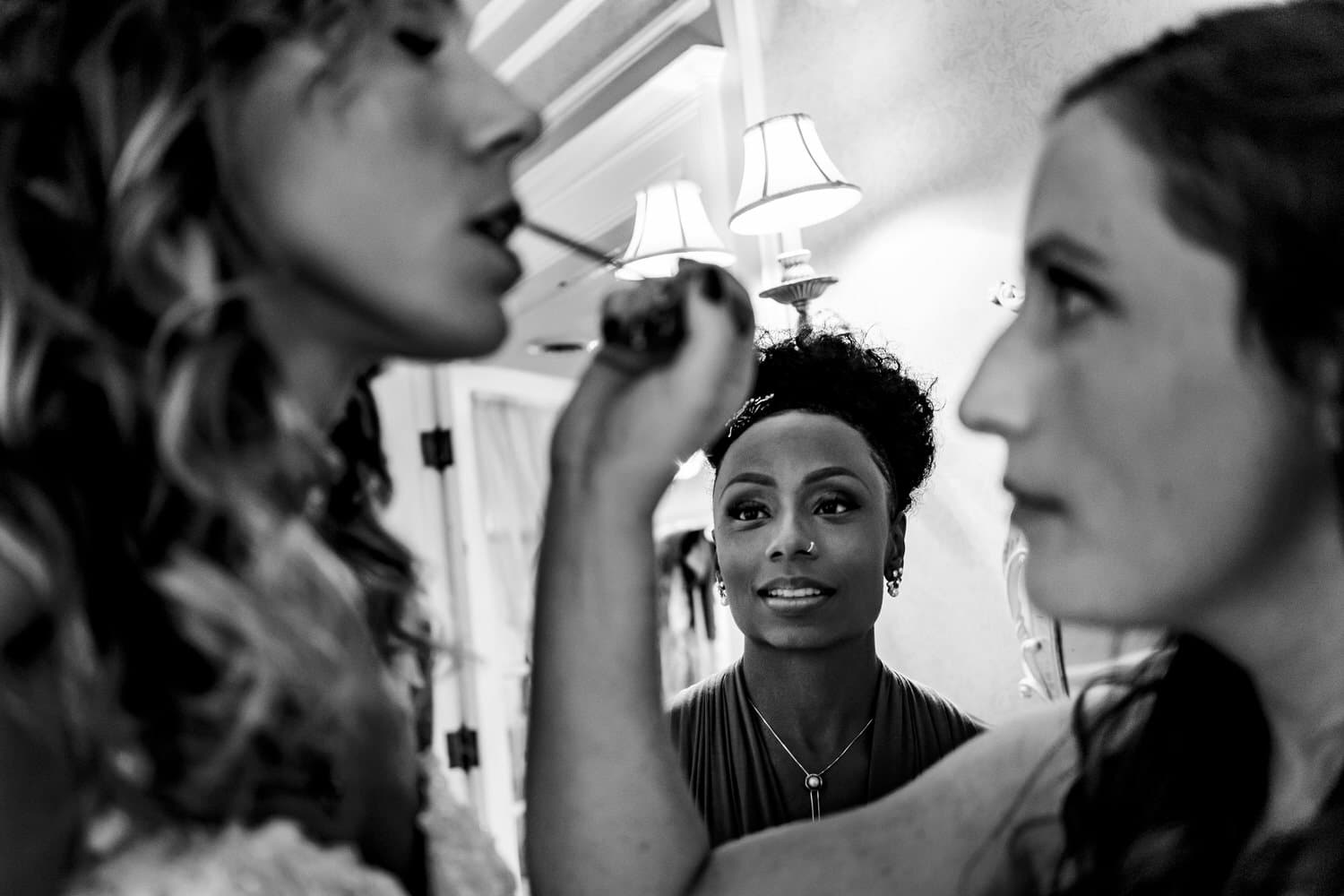 A candid black and white picture of a bridesmaid putting lip gloss onto a bride as a friend watches. 