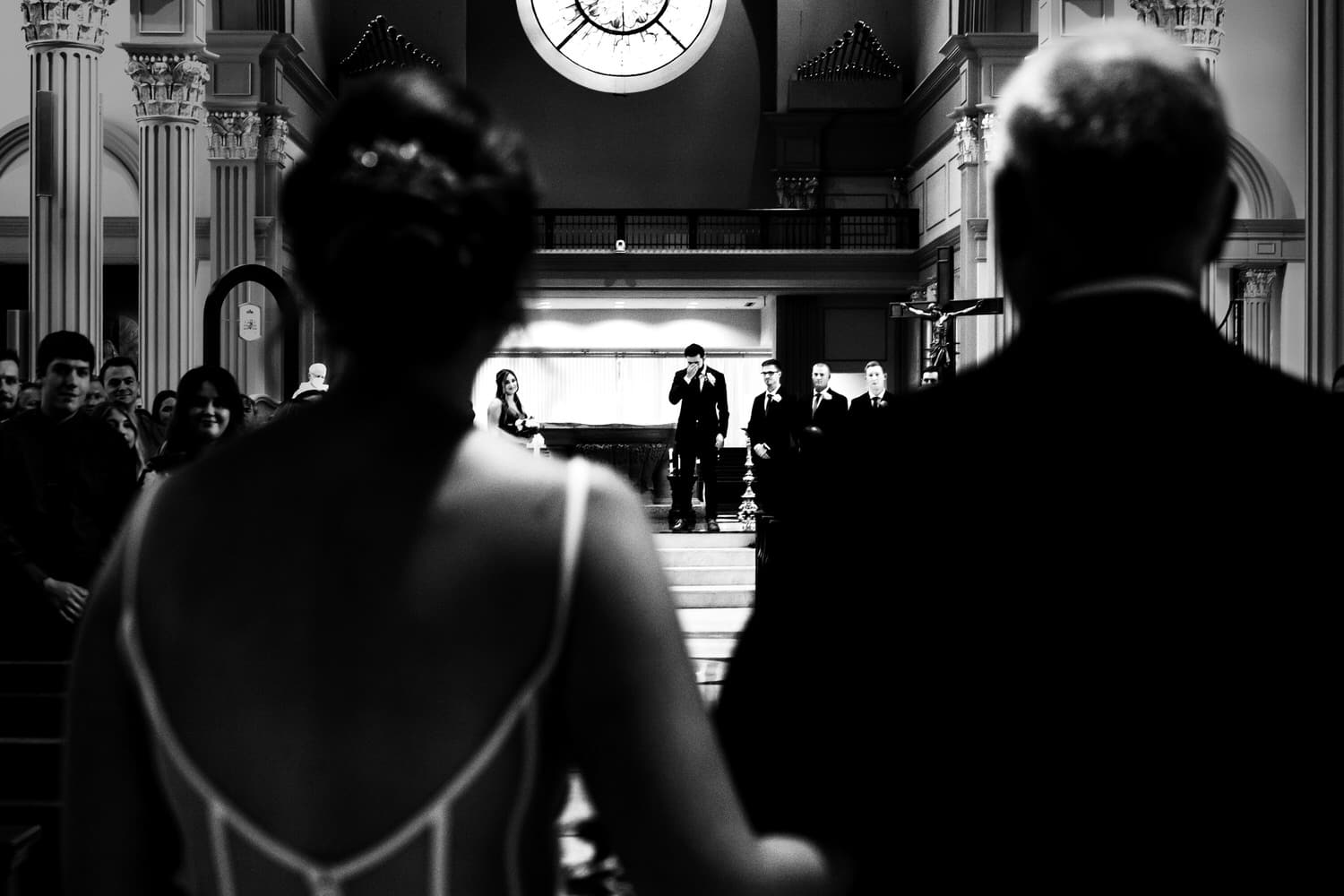 A candid black and white picture of a bride and her dad walking down the aisle of a church, her groom wiping tears from his eyes as she walks towards him on a summer wedding day in Kansas City. 