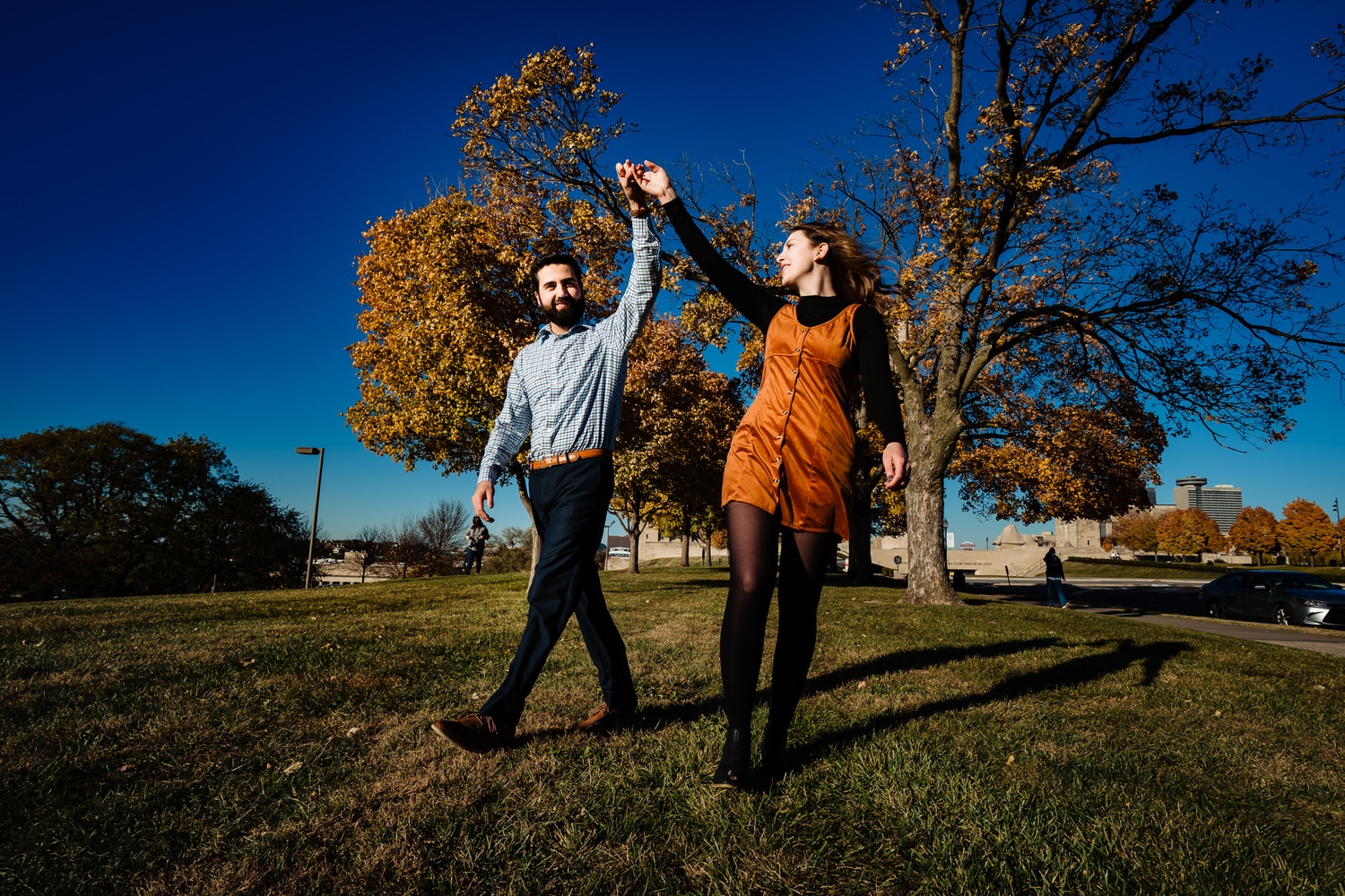 A colorful, candid picture of an engaged couple swinging their held hands up into the air as they walk towards the camera during a fall engagement session at Liberty Memorial in Kansas City. 