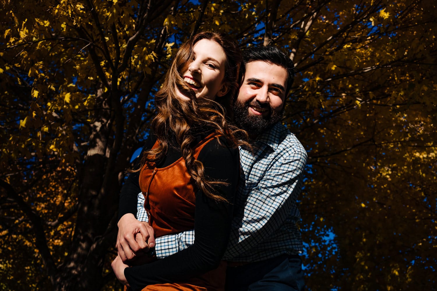 A colorful portrait of an engaged couple wrapped up in an embrace underneath a canopy of bright gold leaves during their colorful downtown KC fall engagement session at Liberty Memorial. 