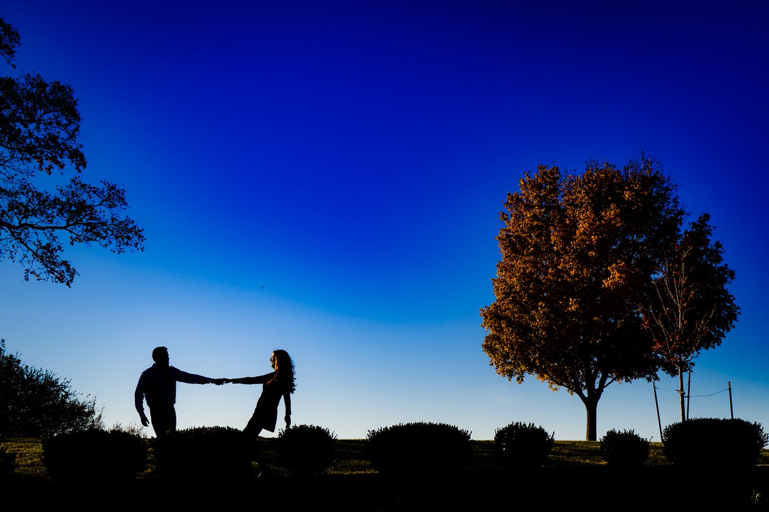 A candid picture of an engaged couple holding hands and leaning apart, silhouetted against a bright blue sky during their Liberty Memorial engagement session in Kansas City. 
