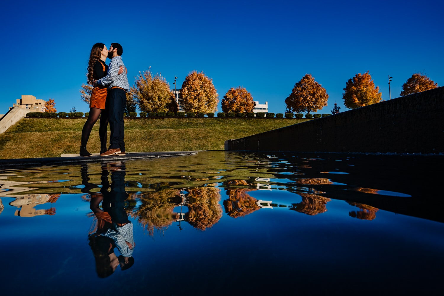 A colorful picture of an engaged couple sharing a kiss, their reflections visible in the water beside them during a fall engagement session at Liberty Memorial. 