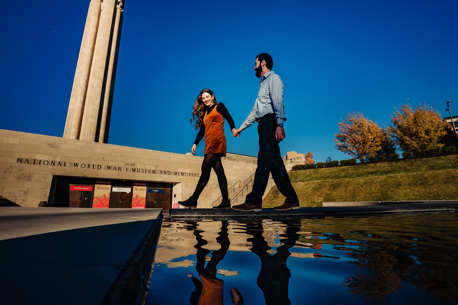 A colorful, candid picture of an engaged couple swinging their held hands up into the air as they walk towards the camera during a fall engagement session at Liberty Memorial in Kansas City. 