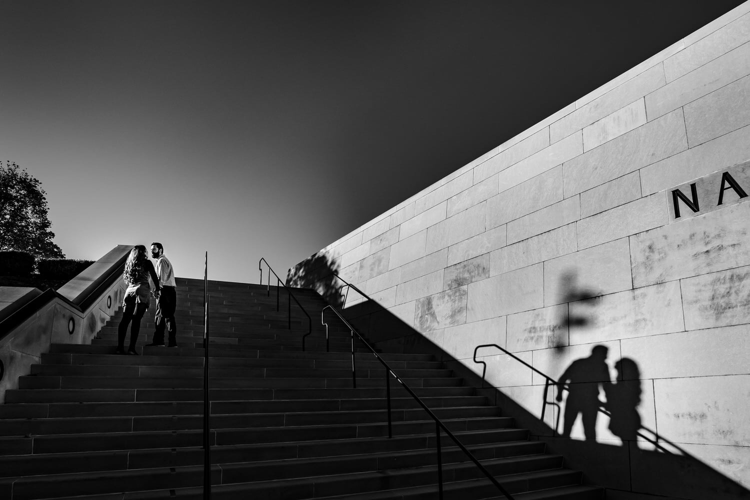 A black and white picture of an engaged couple leaning in to share a kiss as they walk up a set of stairs; their shadows visible on the wall next to them during a fall engagement session in Kansas City. 