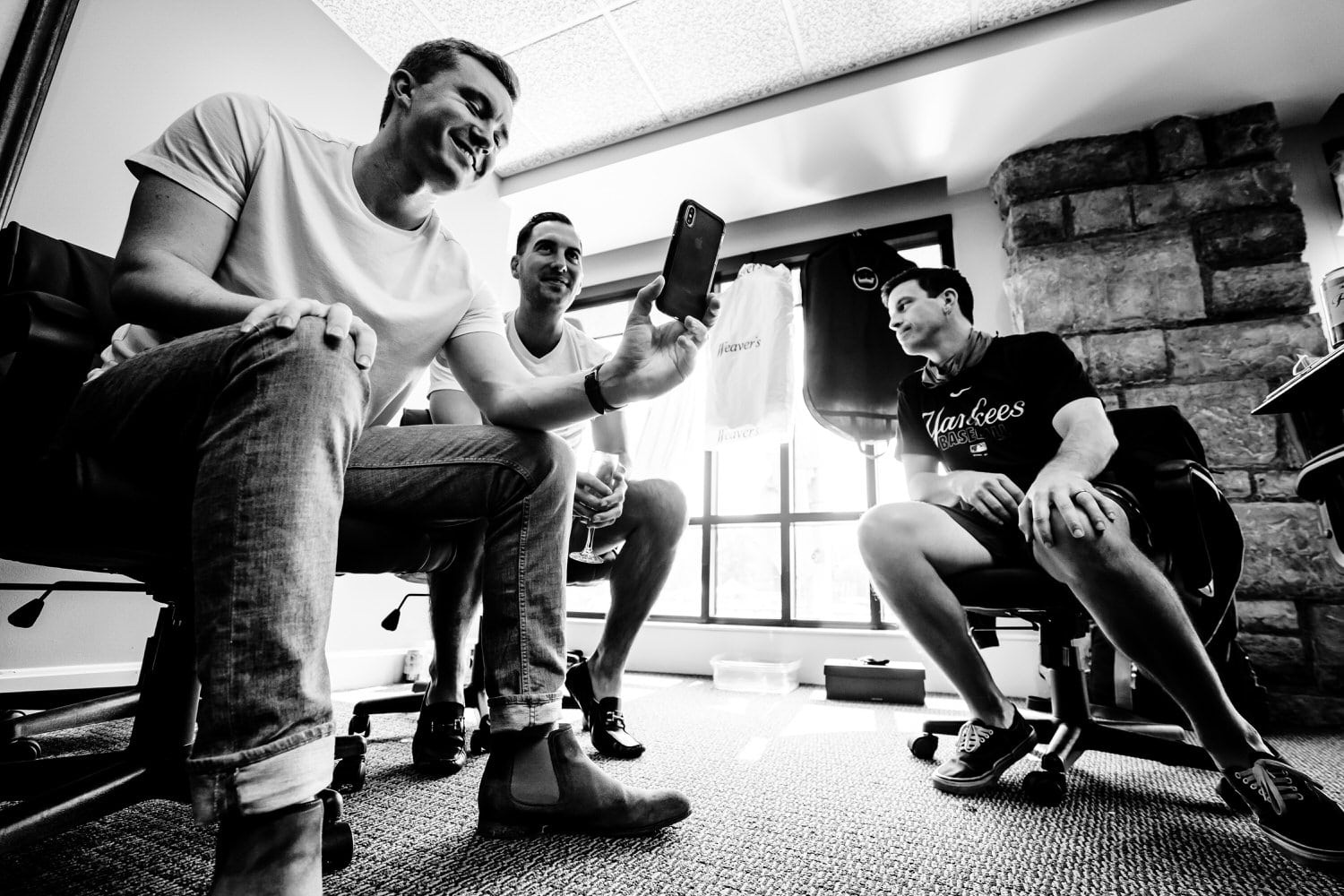 A candid black and white picture of a group of groomsmen laughing at something on a cell phone. 