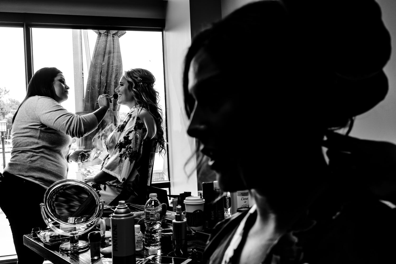 A candid black and white picture of a bride getting her makeup done, a friend getting her hair done visible in the foreground on the morning of a fall wedding at Lake Quivira Country Club in Kansas City. 