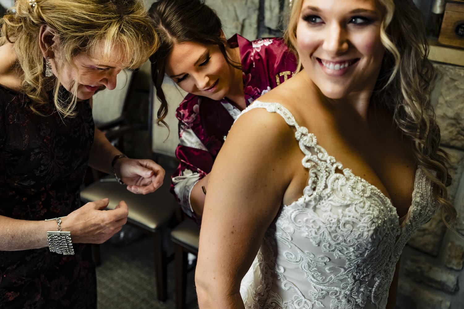 A candid, colorful picture of a woman smiling as her wedding gown gets buttoned on the morning of her wedding. 