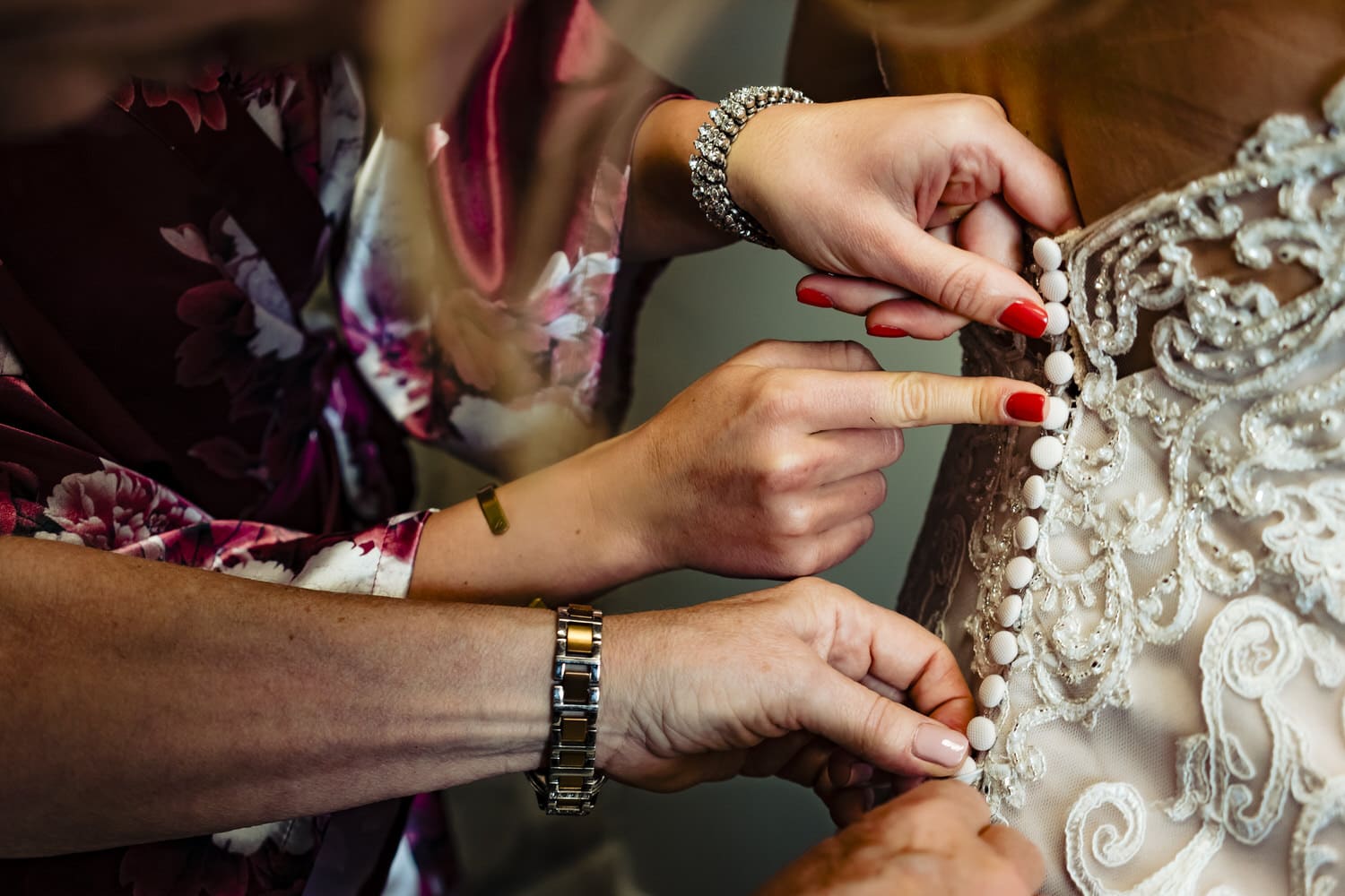 A colorful picture of three sets of hands working together to button the tiny buttons on a wedding gown. 