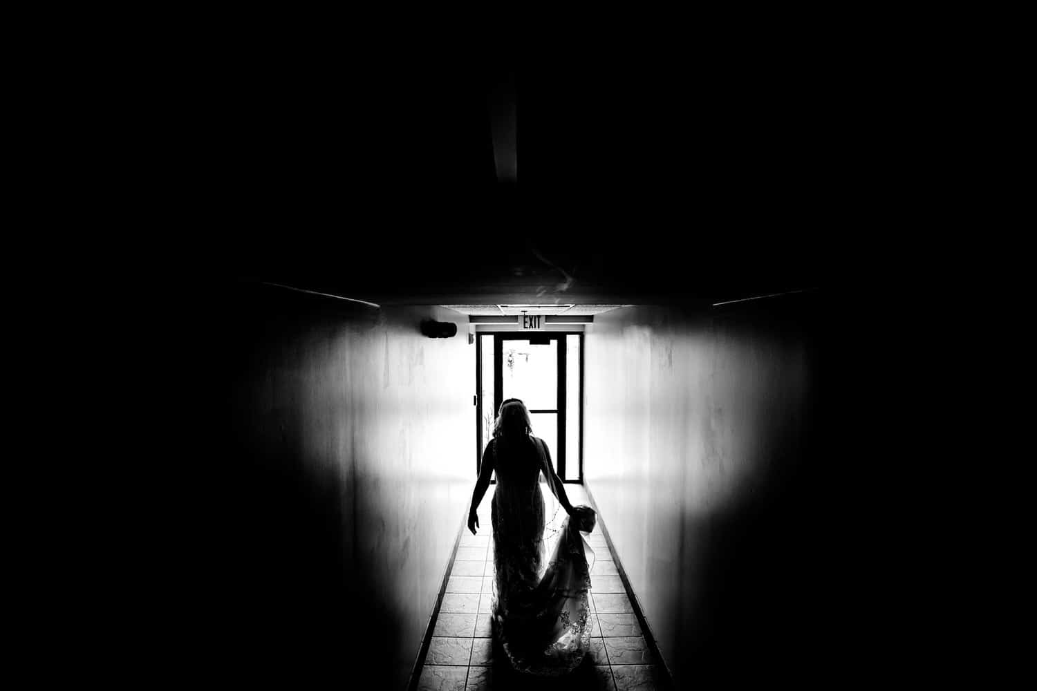 A candid black and white picture of the silhouette of a bride, walking down a long hallway on the day of her fall wedding day. 