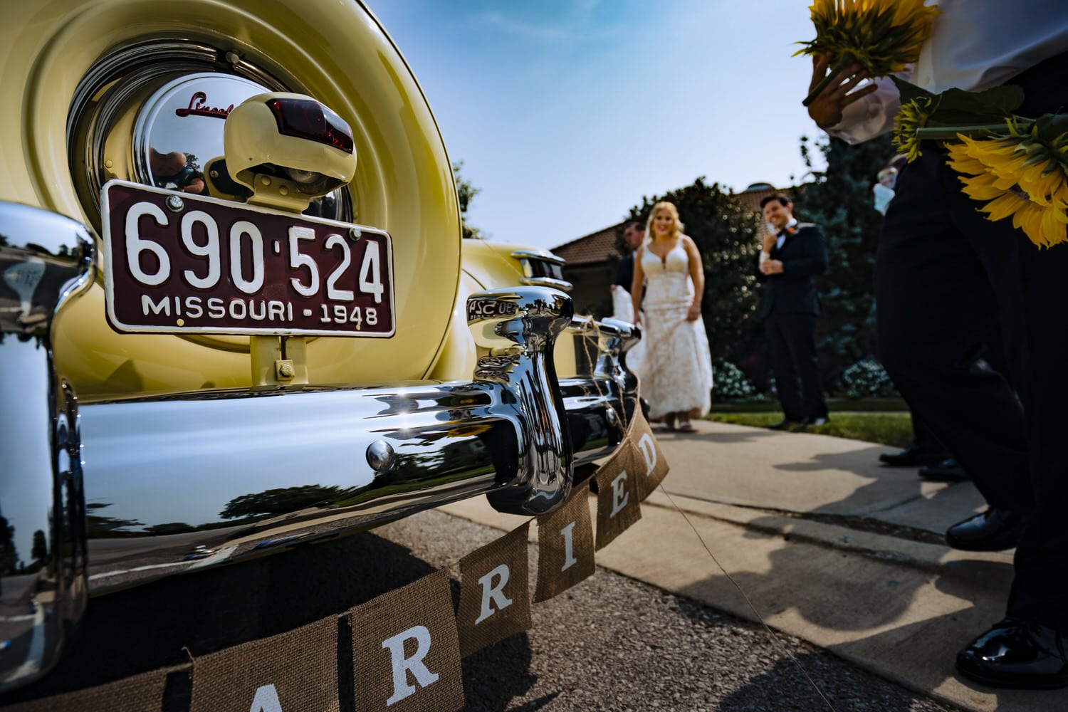 A candid picture of a bride and groom walking up to a vintage yellow car with a "just married" banner attached to the rear bumper on their fall wedding day in Kansas City. 