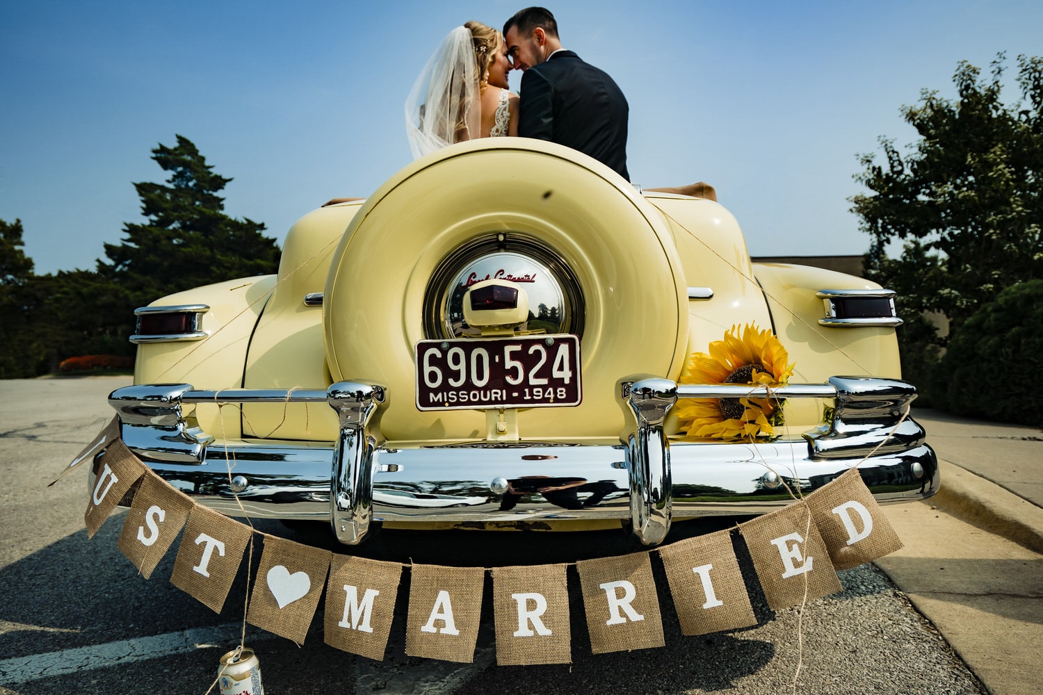 A colorful portrait of a bride and groom sitting in the back of a vintage yellow car, a "just married" banner attached to the back bumper. 