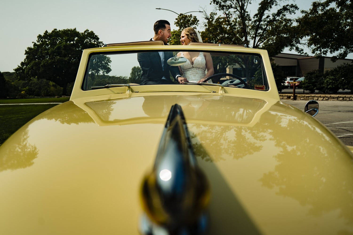 A candid picture of a bride and groom sitting in the back of a yellow vintage convertible, excitedly talking together on their wedding day in Kansas City. 