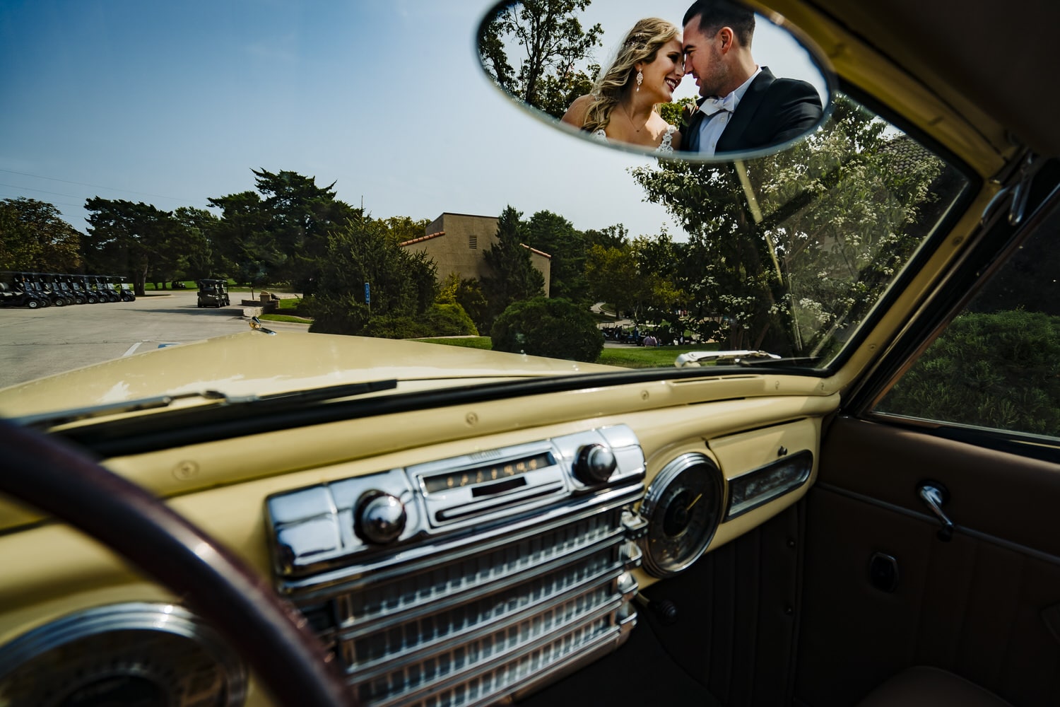 A portrait taken in the rear view mirror of a vintage yellow convertible of a bride and groom leaning in to share a kiss on their fall wedding day at Lake Quivira Country Club. 