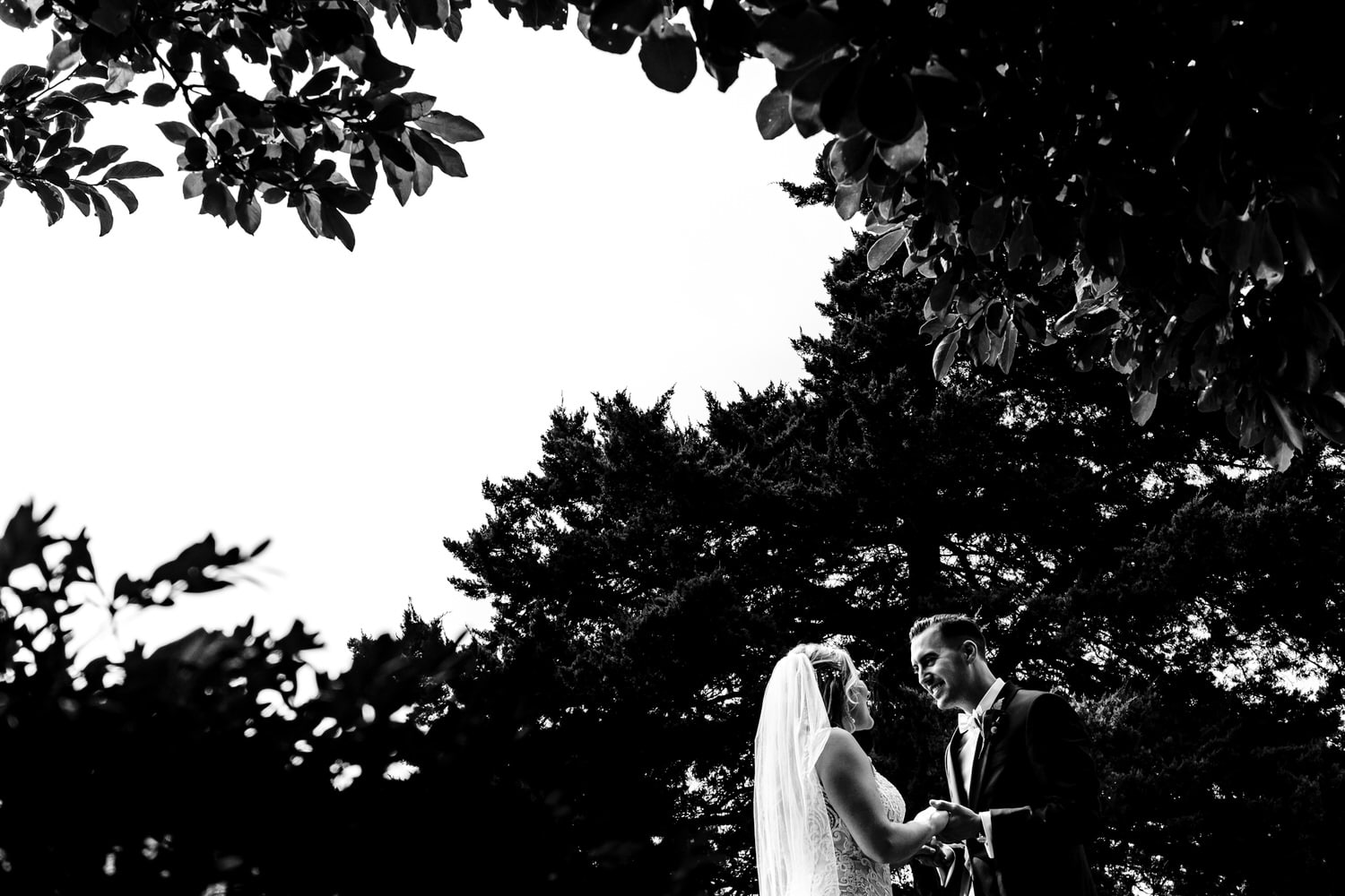 A candid black and white picture of a bride and groom holding hands, laughing together in front of a dark background of pine trees outside Lake Quivira Country Club on their fall wedding day in Kansas City. 