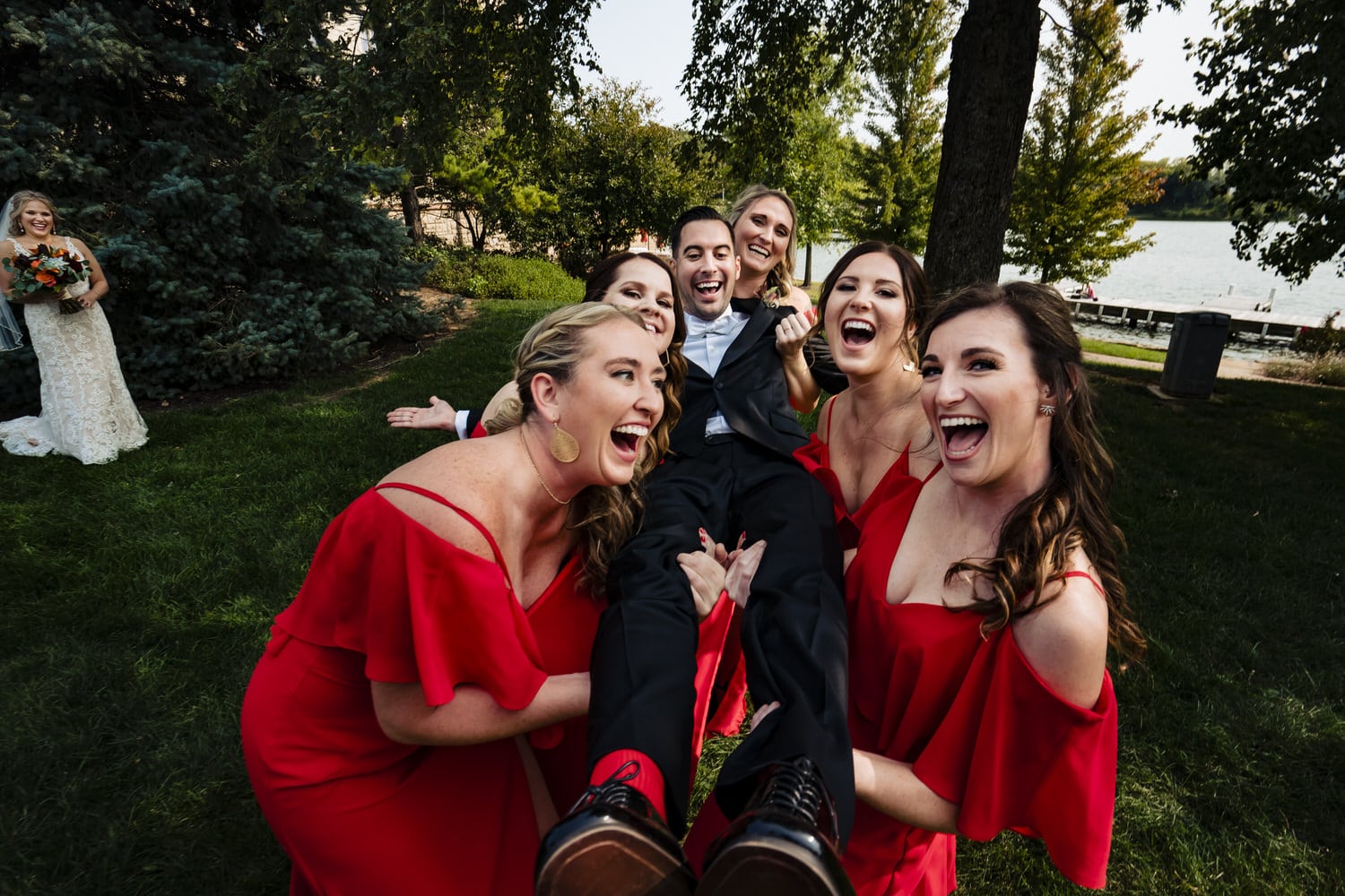 A candid, colorful picture of a group of bridesmaids in red gowns lifting up a groom in a black tuxedo on a fall wedding day in Kansas City. 