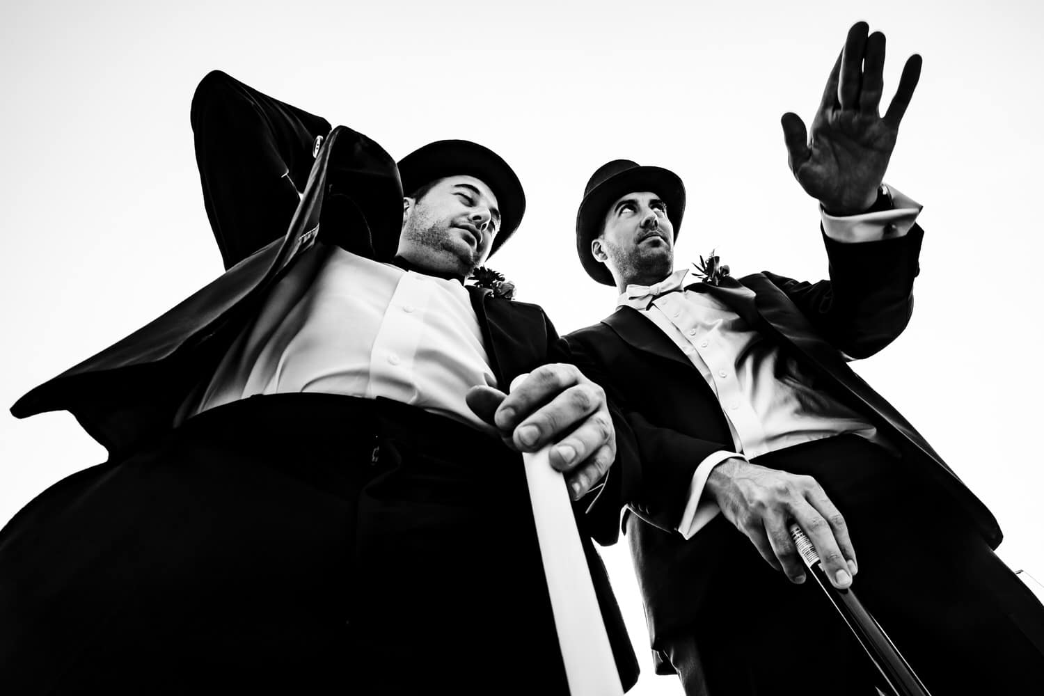 A candid black and white picture taken from the ground looking up of two men in black tuxedos and top hats with canes. 