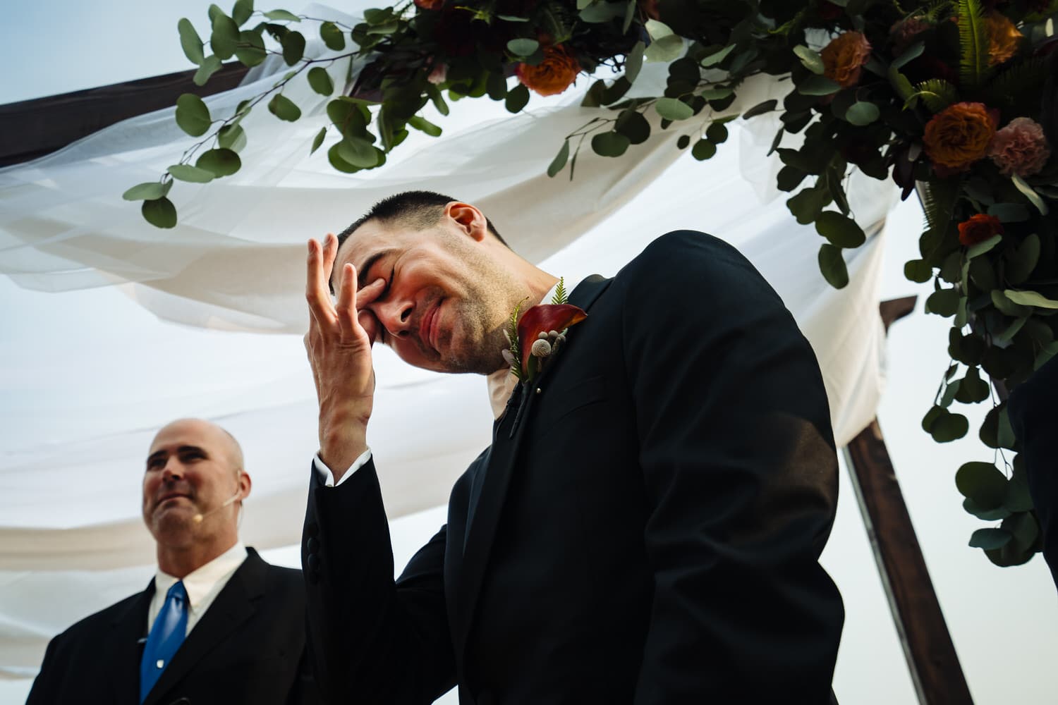 A colorful, candid picture of a groom standing under an arbor, pinching his eyes and trying not to cry as his bride walks down the aisle on a fall wedding day at Lake Quivira Country Club. 