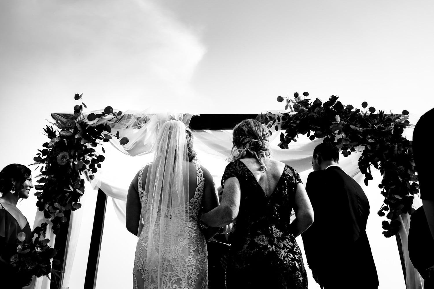 A candid black and white picture of a bride and her mom standing underneath an arbor as the bride is given away during a fall wedding ceremony in Kansas City. 
