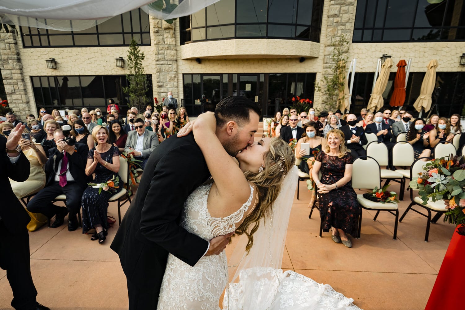 A candid picture of a bride and groom sharing their first kiss, their family and friends celebrating in the background during a fall wedding ceremony at Lake Quivira Country Club in Kansas City. 