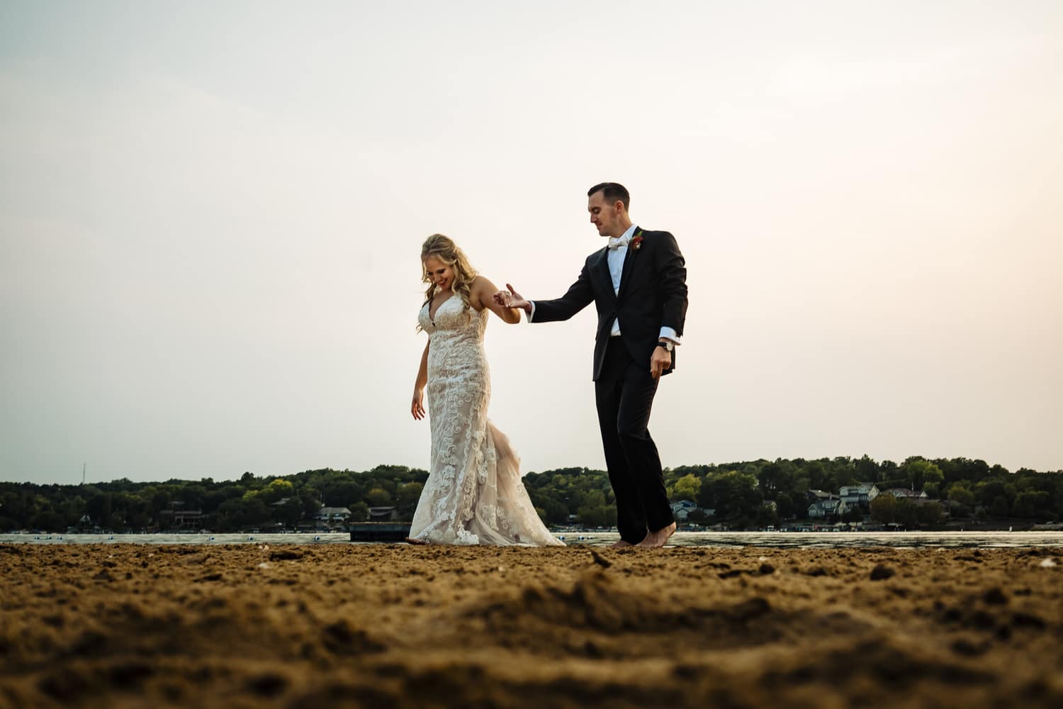 A candid picture of a bride and groom walking barefoot across a beach at Lake Quivira on their fall wedding day in Kansas City. 