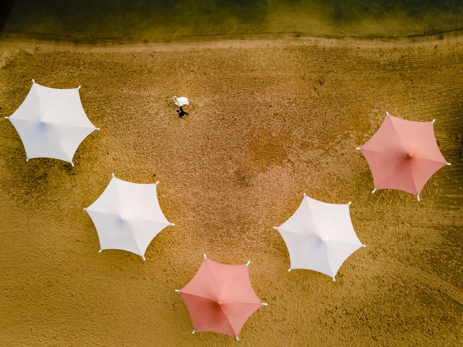 A picture taken by a drone of a bride and groom walking across a beach holding hands, colorful beach umbrellas surrounding them. 