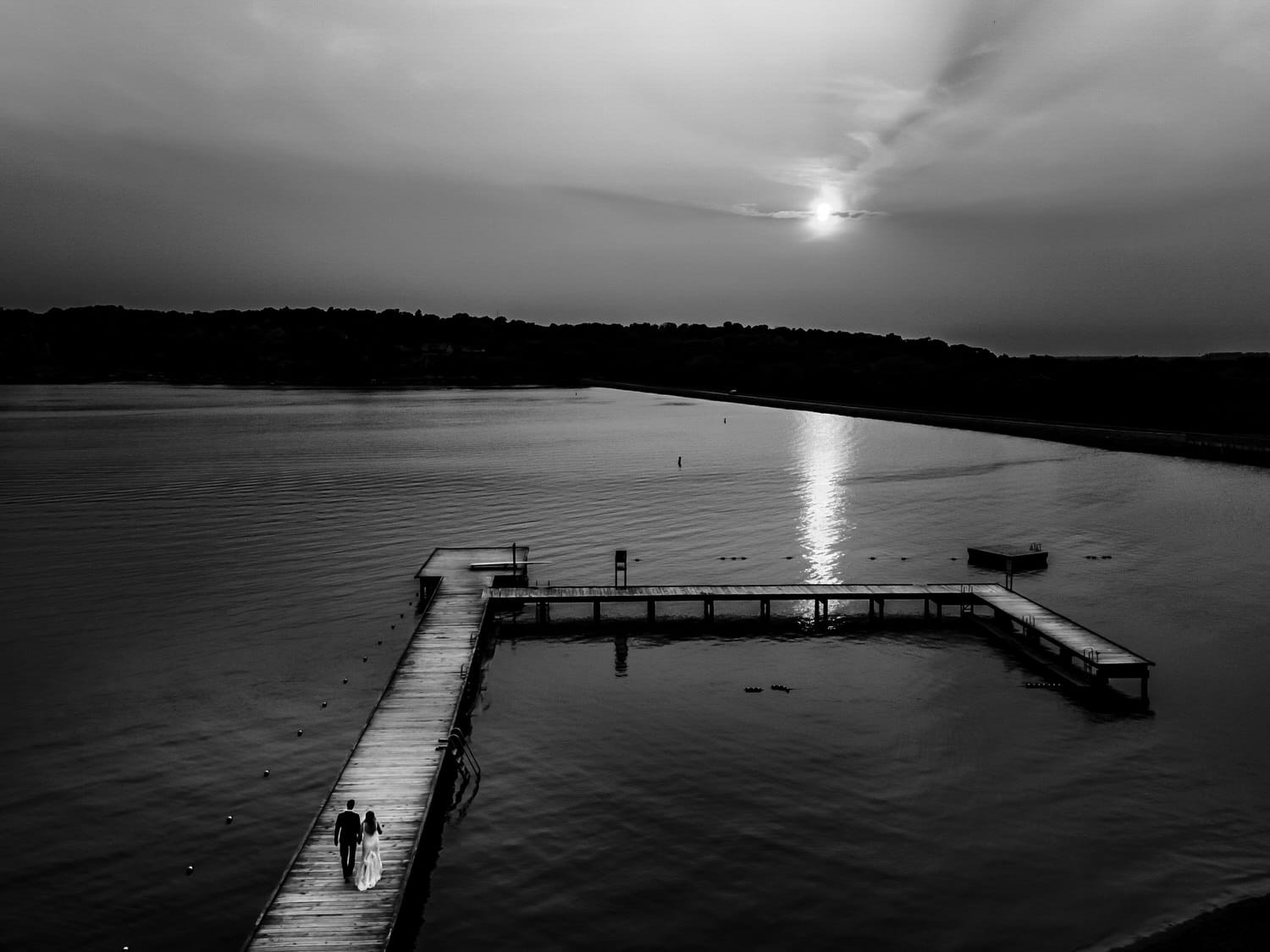 A black and white picture of a bride and groom holding hands walking down the length of a dock extended into Lake Quivira on a fall wedding day in Kansas City. 