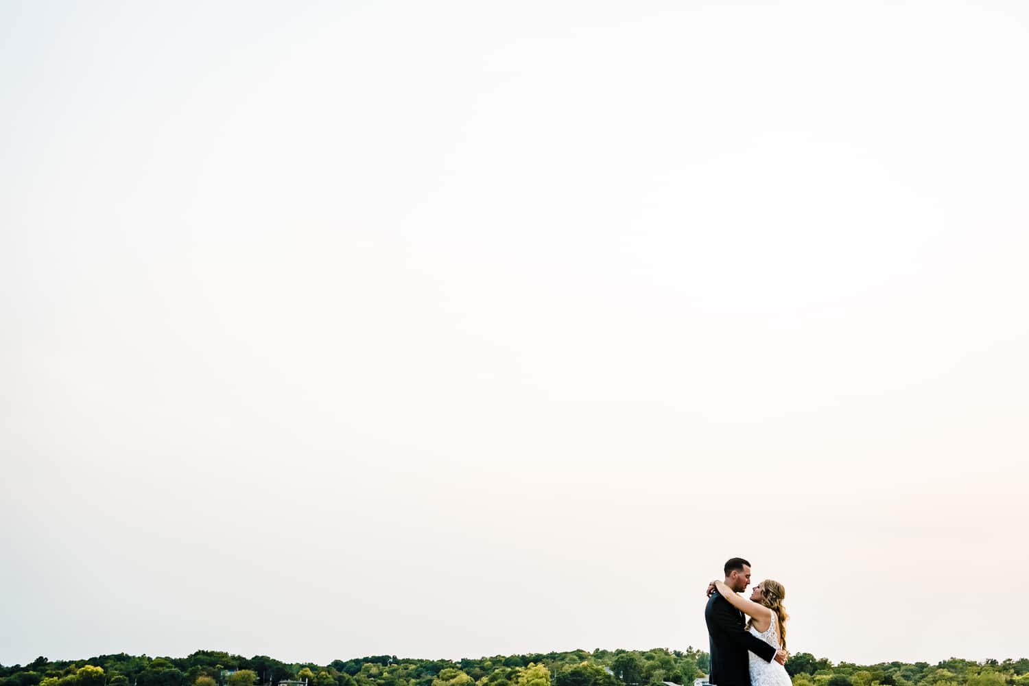A wide, colorful picture of a bride and groom sharing an embrace, hazy skies visible behind them on a fall wedding day in Kansas City at Lake Quivira Country Club. 