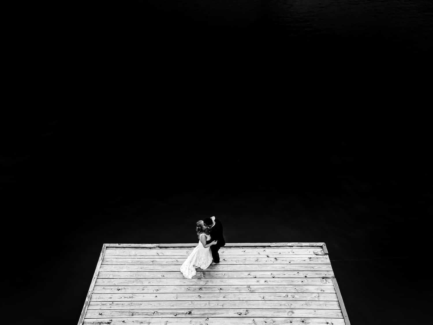 A dramatic black and white portrait of a bride and groom sharing a kiss as they stand on the edge of a dock on their wedding day. 
