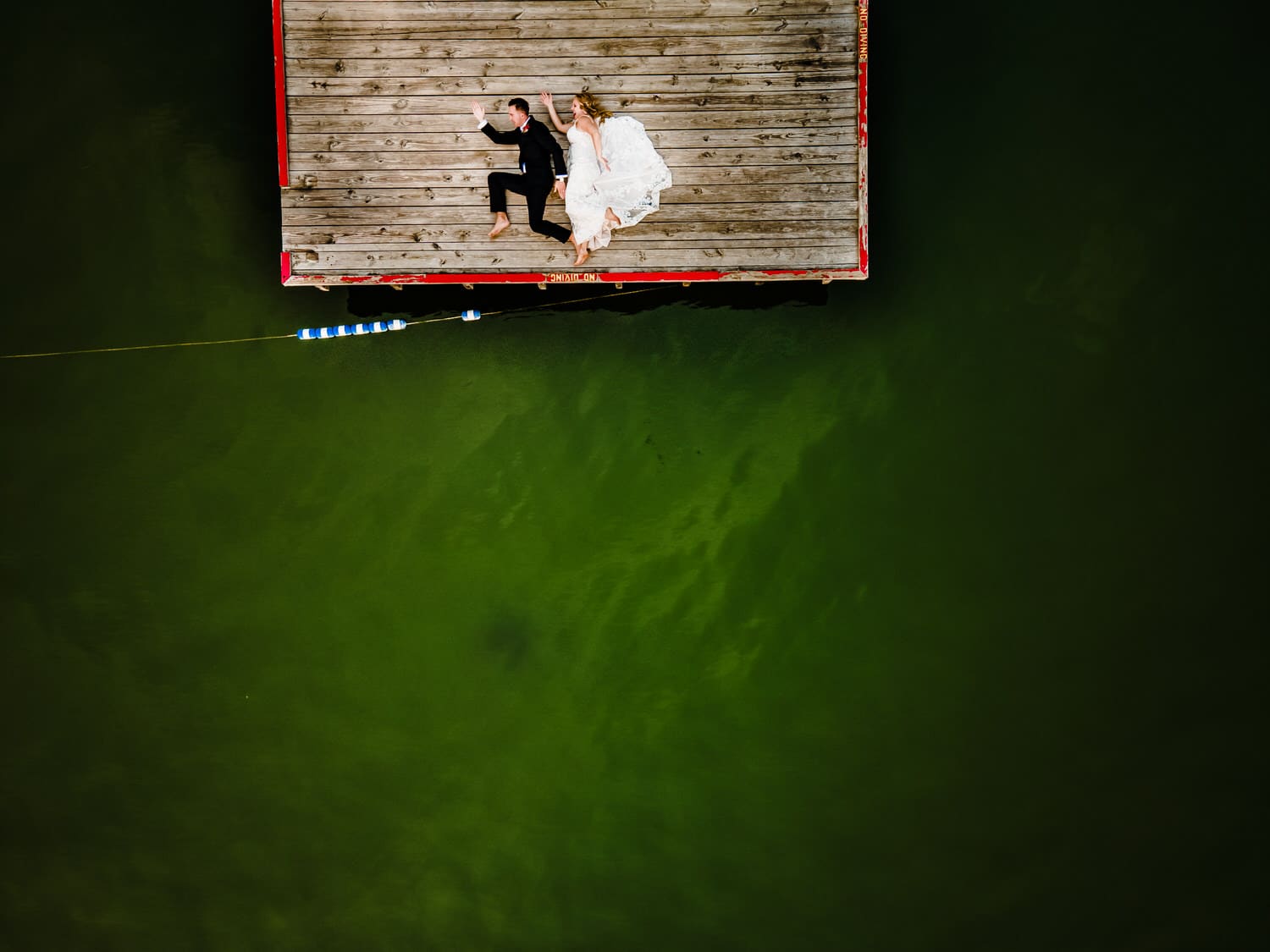 A picture taken by a drone of a bride and groom laying on a dock, their bodies in a running motion on their wedding day at Lake Quivira Country Club in Kansas City. 