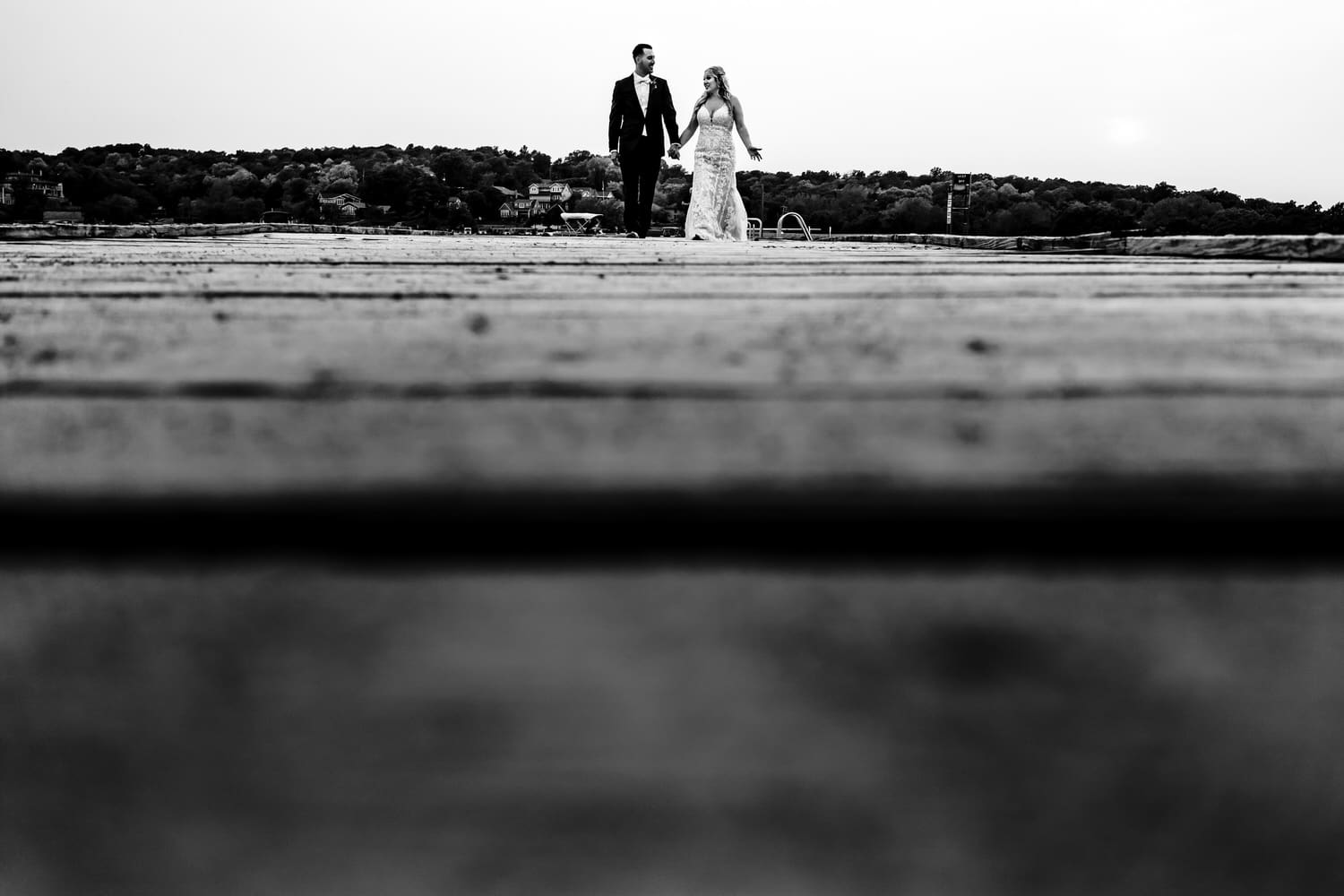 A candid black and white picture of a bride and groom holding hands walking down a dock on their fall wedding day at Lake Quivira Country Club in Kansas City. 