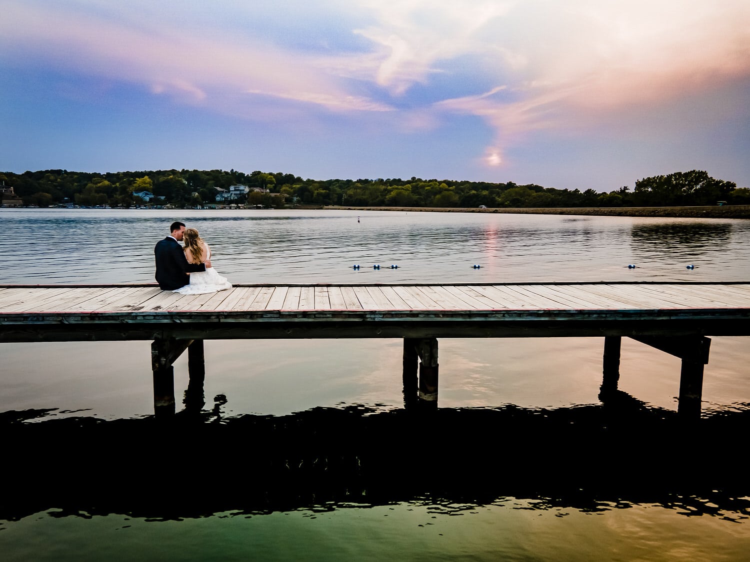 A colorful picture of a bride and groom sitting on a dock at sunset on their fall wedding day at Lake Quivira Country Club in Kansas City. 