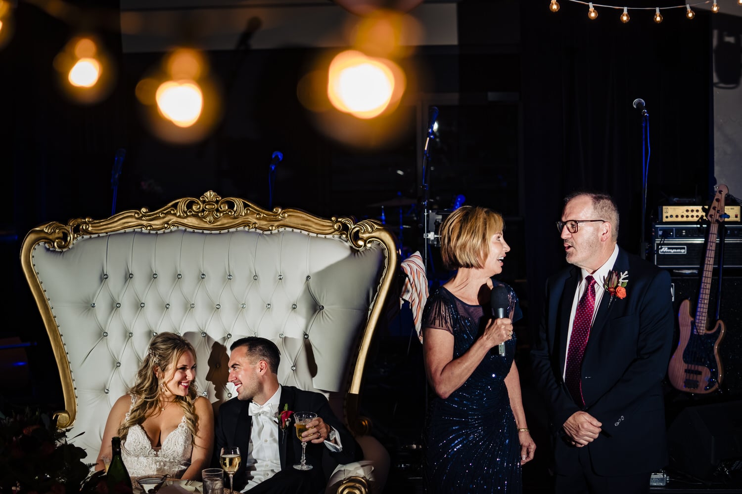 A candid picture of a bride and groom sitting in a large chair, smiling at each other as his parents give a toast during a wedding reception at Lake Quivira Country Club in Kansas City. 