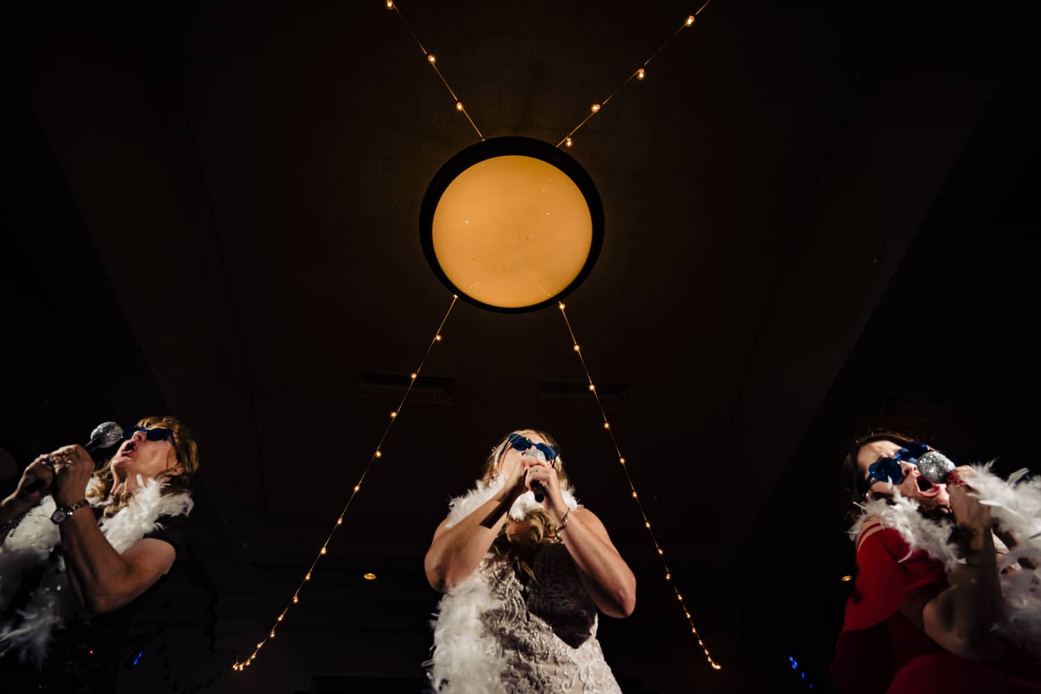 A candid picture taken from the ground looking up of three women in feather boas singing a song with faux microphones during a wedding reception at Lake Quivira Country Club in Kansas City. 