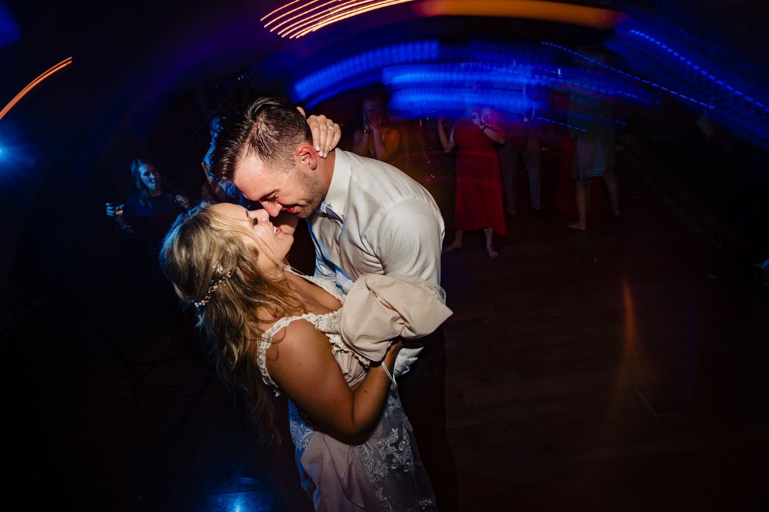 A colorful, candid picture of a bride and groom sharing a kiss in the middle of a crowded dance floor during their wedding reception at Lake Quivira Country Club in Kansas City. 