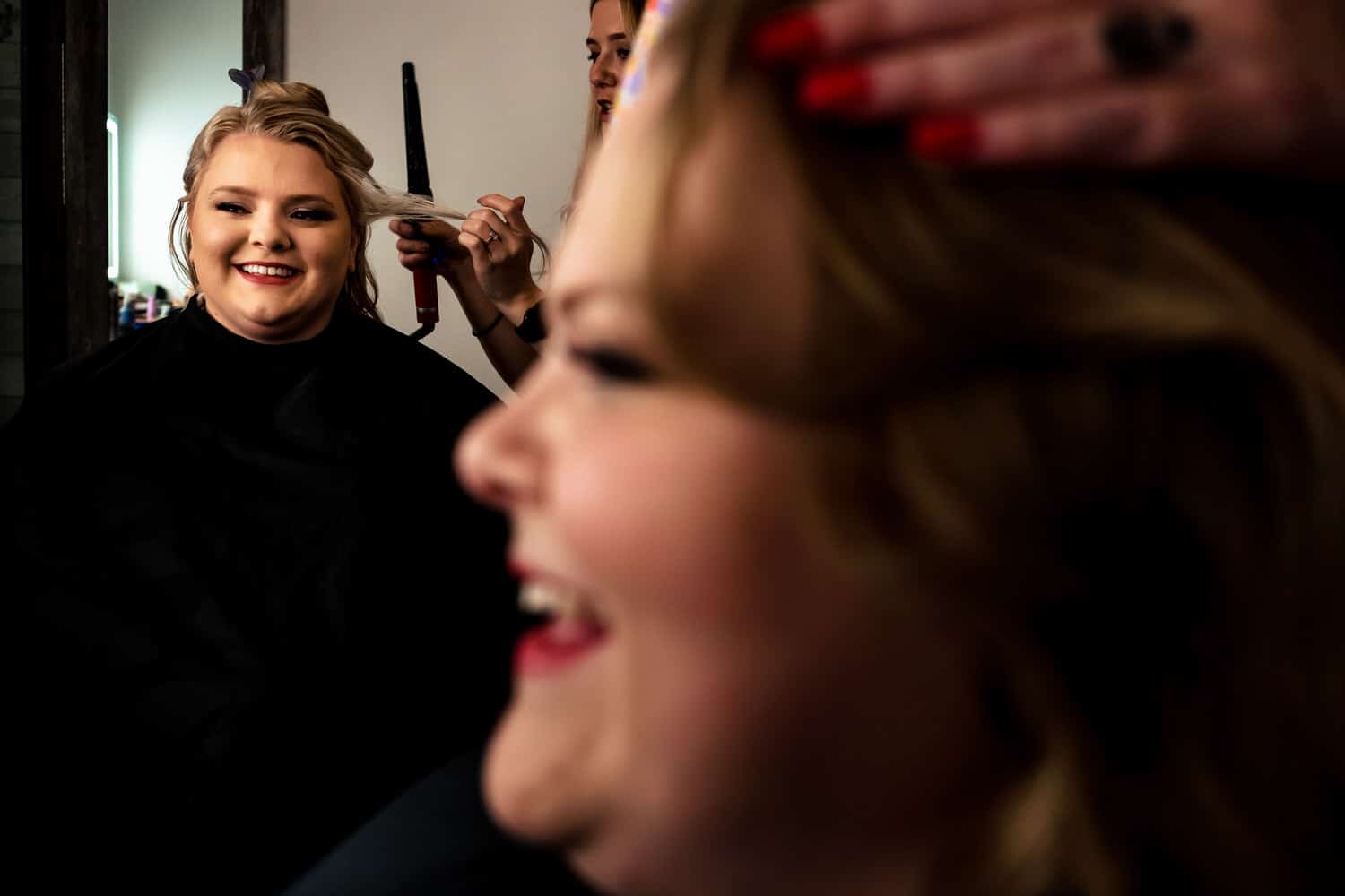 A candid, colorful picture of a woman getting her hair curled in the background, as her sister laughs candidly in the foreground. 