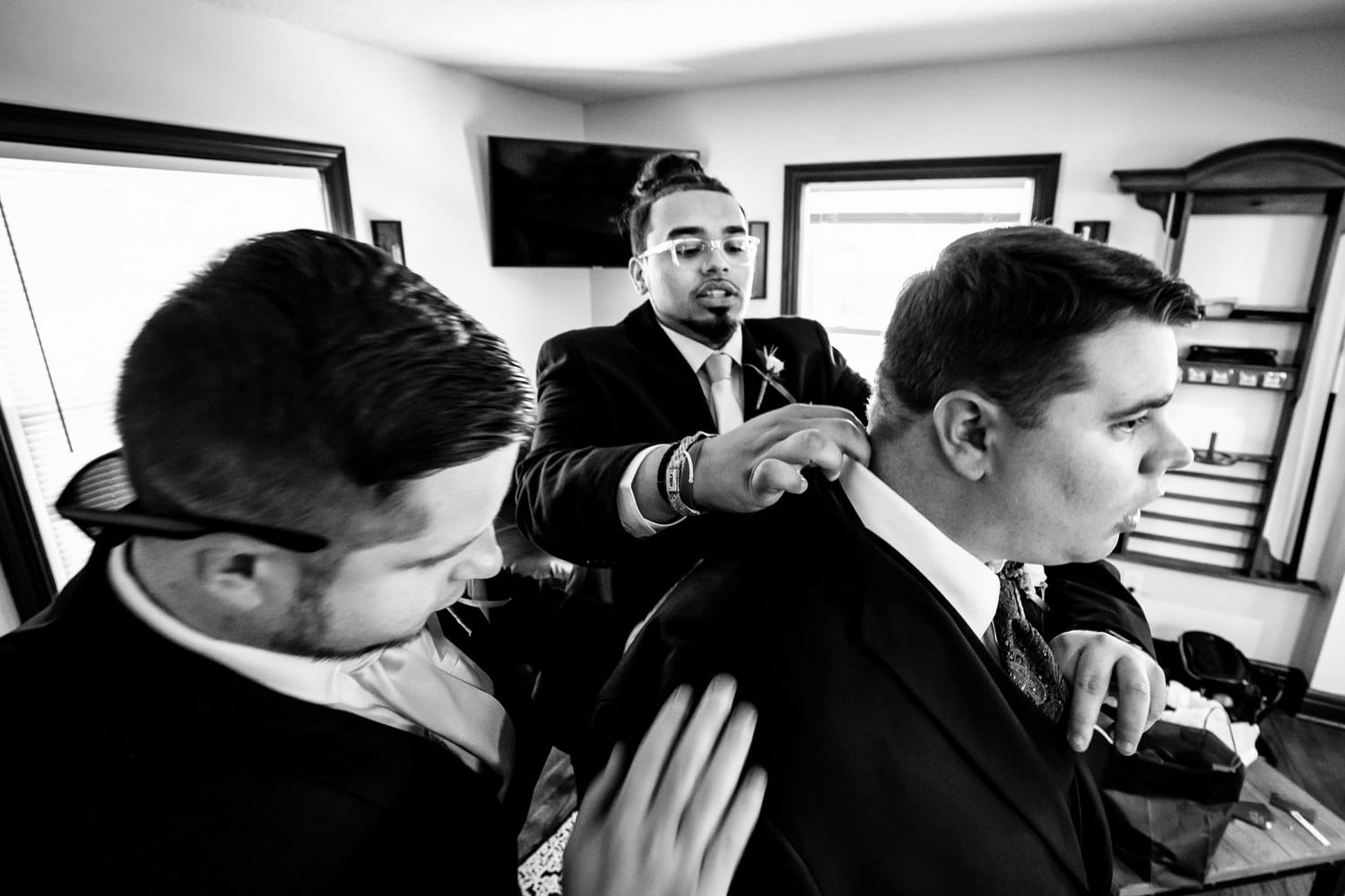 A candid black and white picture of a group of groomsmen helping a groom make last minute adjustments to his tux on his wedding day. 