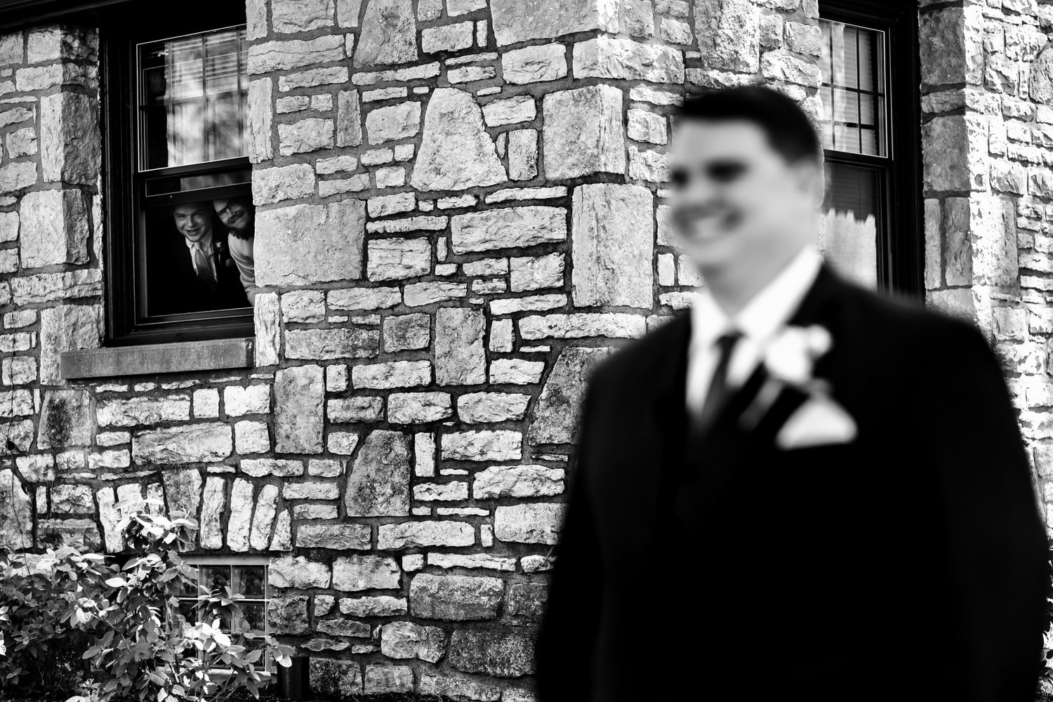A candid black and white picture of a groom waiting for his bride, his groomsmen peeking out of a nearby window. 