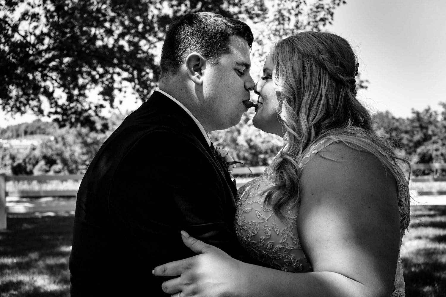 A candid black and white picture of a bride and groom sticking their tongues out at each other, with just the tips touching on their summer wedding day. 